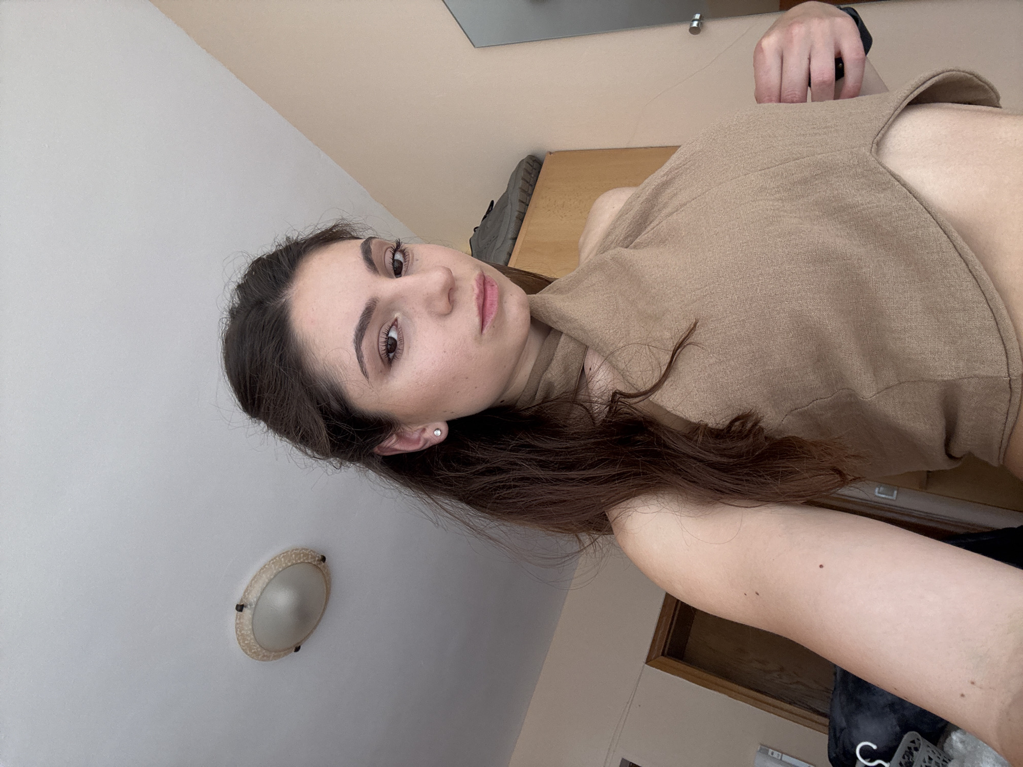 In a medium shot, a young woman with long, dark brown hair is captured in what appears to be a bedroom. She is wearing a tan, halter-neck top. The background reveals a light-colored wall, a ceiling light fixture, a wooden closet with a shoe on top, and a doorway. The overall ambiance suggests an indoor, domestic setting, likely in Varna, Bulgaria, given the context provided. The woman is looking directly at the camera with a neutral expression.