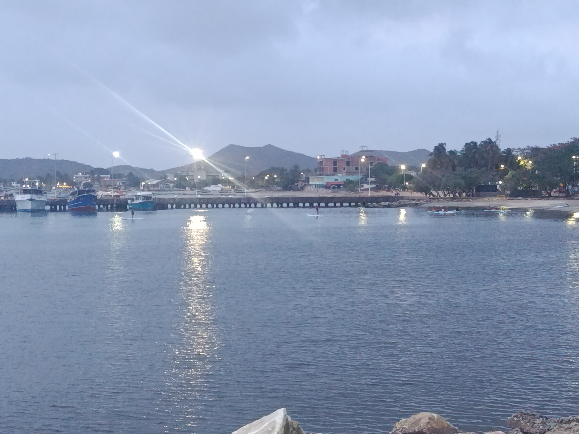 The image depicts the harbor in Juan Griego, Venezuela, at dusk. The water is calm and reflects light from the street lamps lining the shore and pier. Several boats are docked on the left side of the harbor. A pier extends into the water, and two people on paddleboards are visible. The shoreline features a sandy beach with scattered trees and buildings in the background. Mountains are visible in the distance. In the foreground, rocks line the shore, indicating the edge of the harbor.
