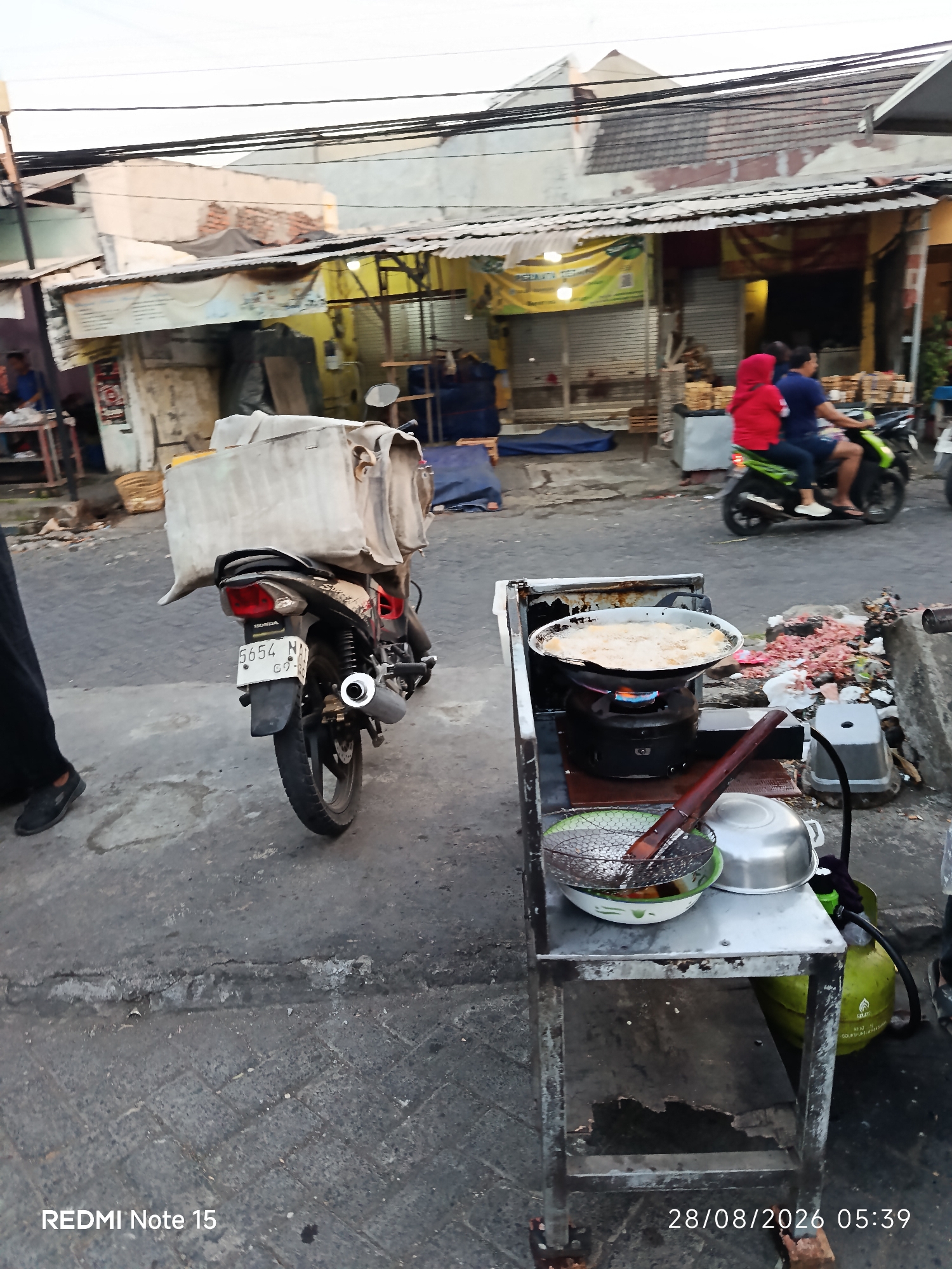 The image depicts a street scene in Keboansikep, Indonesia, likely in the late afternoon or early evening given the lighting. In the foreground, a street food vendor's cart is set up on a paved street. A wok, filled with oil and food being fried, is positioned on a portable stove. Various cooking utensils, including a metal strainer and a lidded pot, are on the cart. A propane tank is visible beneath the cart. Behind the cart, a motorcycle with a large, covered cargo box is parked. The motorcycle has a license plate with the number "5654 N". In the background, buildings with corrugated metal roofs line the street. Two people are visible riding a motorcycle in the distance, with the person in the back wearing a red hijab. The environment appears to be a busy, commercial area with a slightly cluttered and weathered aesthetic.