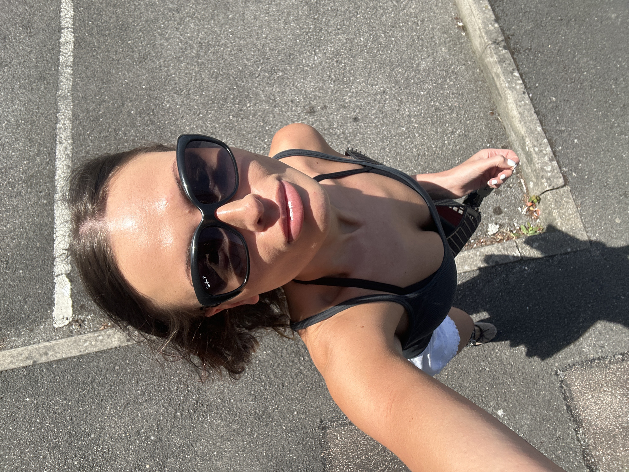 A high-angle, medium shot captures a woman in her late 20s or early 30s from the chest up, posing for a selfie outdoors on a paved surface, possibly a parking lot or sidewalk in Tunbridge Wells, United Kingdom, under bright sunlight that casts distinct shadows. She has medium-length, dark brown hair that is slightly windblown and she is wearing large, dark-rimmed sunglasses. Her lips are slightly parted, and she appears to be looking directly into the camera. She is wearing a dark, ribbed tank top with thin straps, revealing her shoulders and upper chest. A white skirt is visible beneath the tank top. A dark-colored handbag with a patterned strap is slung over her left shoulder, and her right arm is extended, holding what appears to be a mobile phone, out of frame. Her left hand is visible near the bottom of the frame, resting on her lap, with neatly manicured white-painted fingernails. The background consists of a textured asphalt surface with a distinct white painted line, suggesting a marked area such as a parking space or road marking. A concrete curb runs along the right side of the frame, with some dried grass and debris near its base. The overall impression is a casual, outdoor snapshot taken on a sunny day.