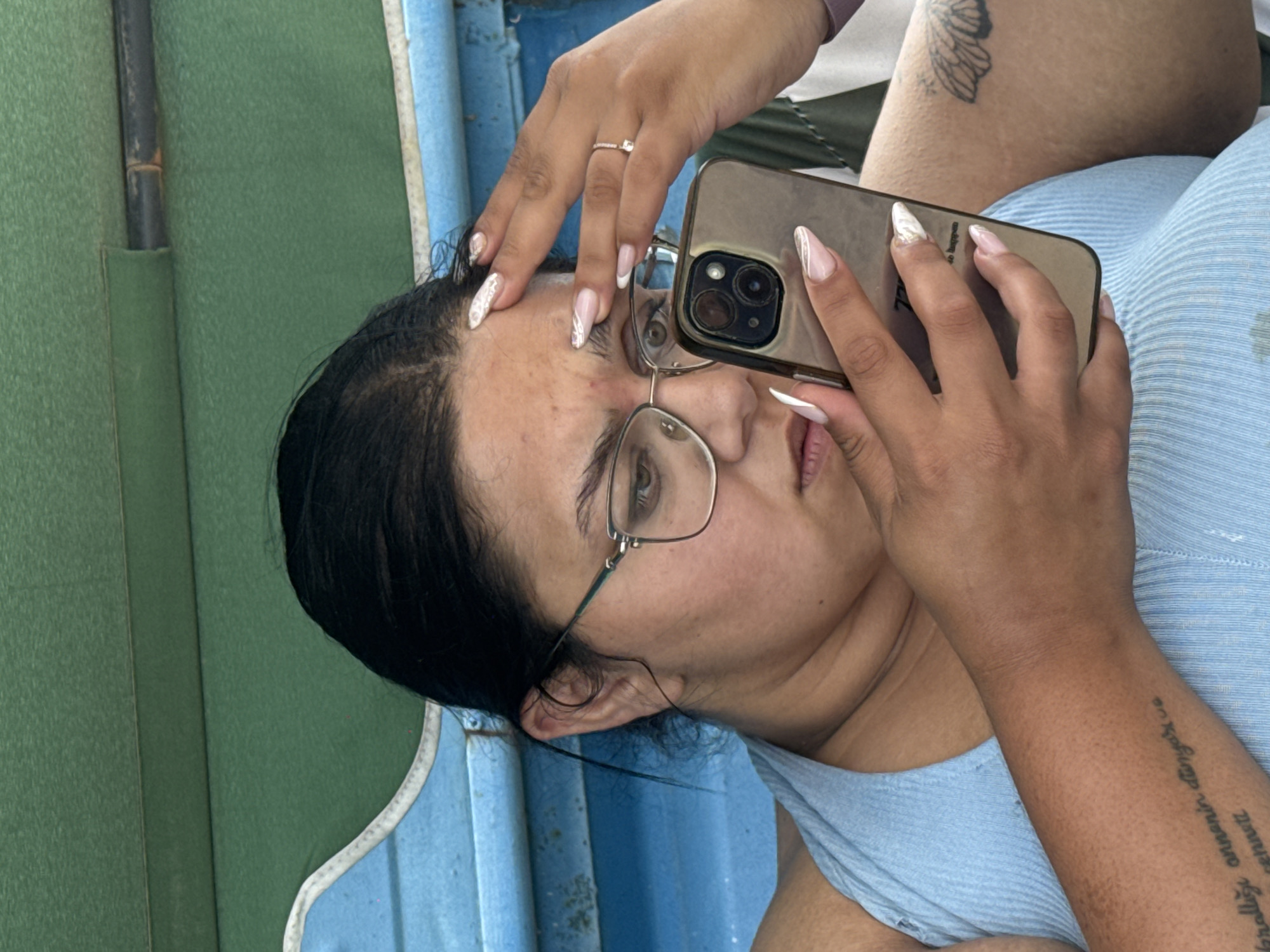 A young woman with dark hair and glasses is seated indoors, possibly in a boat, given the blue and green upholstery. She is looking down at her phone, which she is holding in her right hand. Her left hand is resting on her forehead. She is wearing a light blue tank top. There is a tattoo on her left forearm and another on her right thigh. The background shows textured green and blue surfaces. The image does not contain any information that could place it in the city of Sredets, Bulgaria.