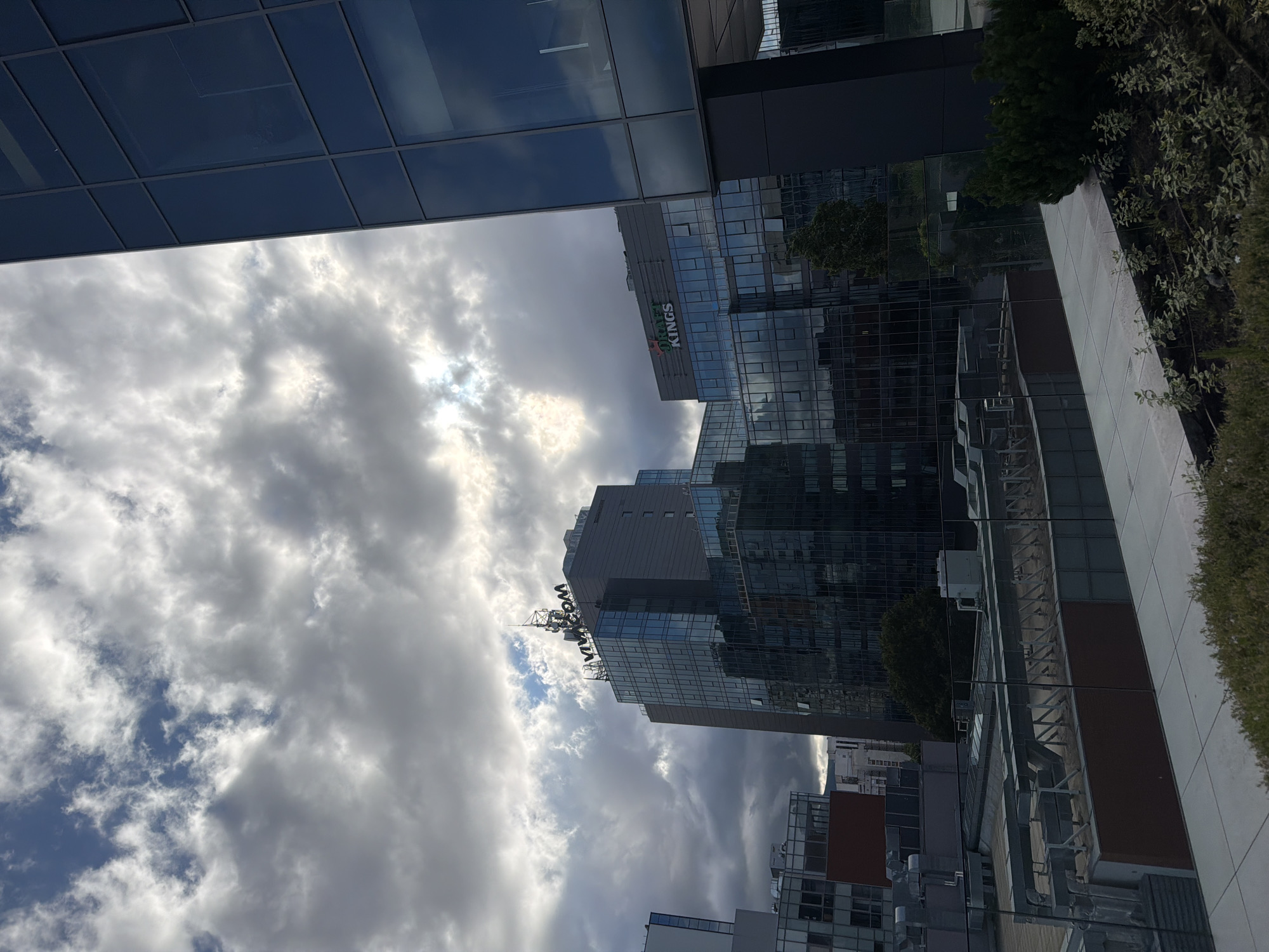 The image displays a modern cityscape under a cloudy sky, captured from a slightly elevated perspective, possibly from a lower building or balcony. The scene is dominated by several contemporary buildings, characterized by their glass and concrete facades.
In the foreground and to the left, a dark, reflective glass-paneled building partially obscures the view. In the midground, a prominent high-rise building features extensive glass exteriors, reflecting the sky and other structures. On this building, green letters clearly spell "PLAYTECH KINGS." Further into the background, another tall, glass-clad building is visible, and atop its roof structure, the word "VIVACOM" is displayed in white, inverted text, surrounded by antennas.
The sky is mostly covered with large, white and gray cumulus clouds, with the sun breaking through in the upper part of the frame, creating bright, diffused light. Patches of blue sky are visible between the clouds. On the right side of the image, what appears to be a flat rooftop or terrace area is lined with neatly trimmed green hedges and shrubs.
The time of day appears to be daytime, with bright but diffused sunlight due to the cloud cover. The overall setting is an urban business or commercial district in Sofia, Bulgaria, characterized by modern architecture and a dynamic sky. There is no specific activity taking place; the image is a static architectural and sky scape.