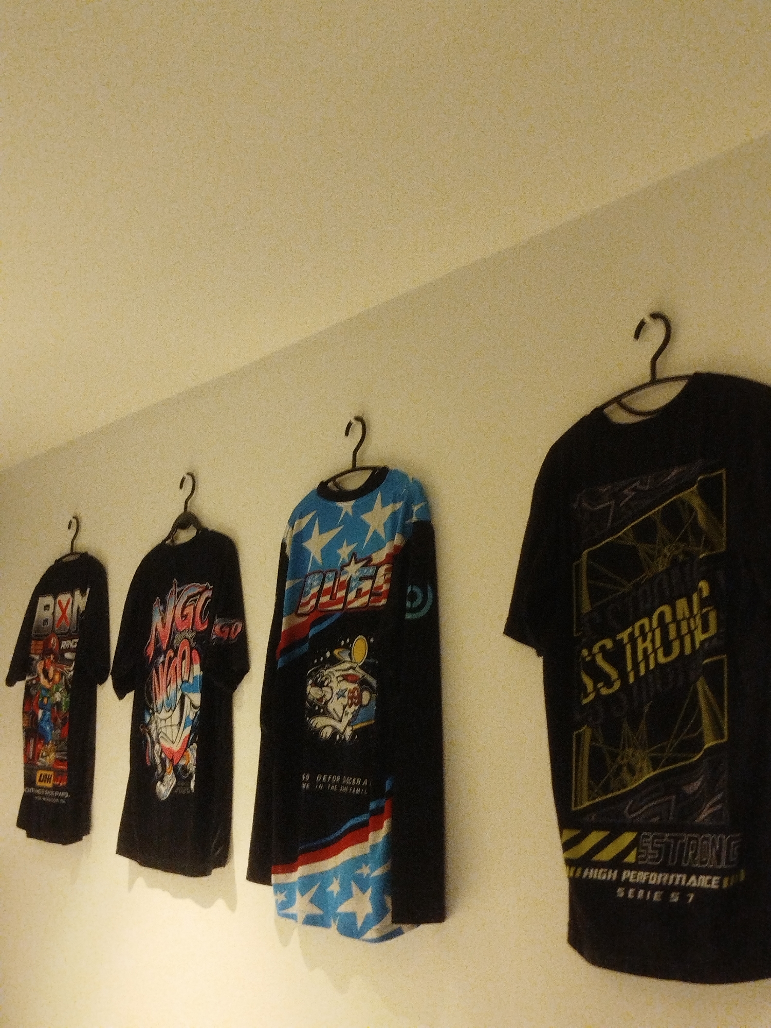 Four black t-shirts are displayed on hangers against a white wall. The t-shirts have various graphics and text printed on them. The leftmost shirt features a colorful graphic with text that appears to say "ORIGE MOS PARS" and "RING". The second shirt from the left has a graphic with the text "NGC" in large, stylized letters, with red and blue accents. The third shirt, a long-sleeved one, has a design with blue and white stars, red and blue stripes, and text that reads "19 DEFOR SCARA WIN THE SHEFAMIL". The rightmost shirt has a graphic with yellow accents and the text "SSTRONG" and "55TRONE" along with "HIGH PERFORMANCE SPA SERIES 7". The lighting suggests an indoor setting, possibly a shop or a personal collection display. The overall mood is casual and focuses on the apparel.
