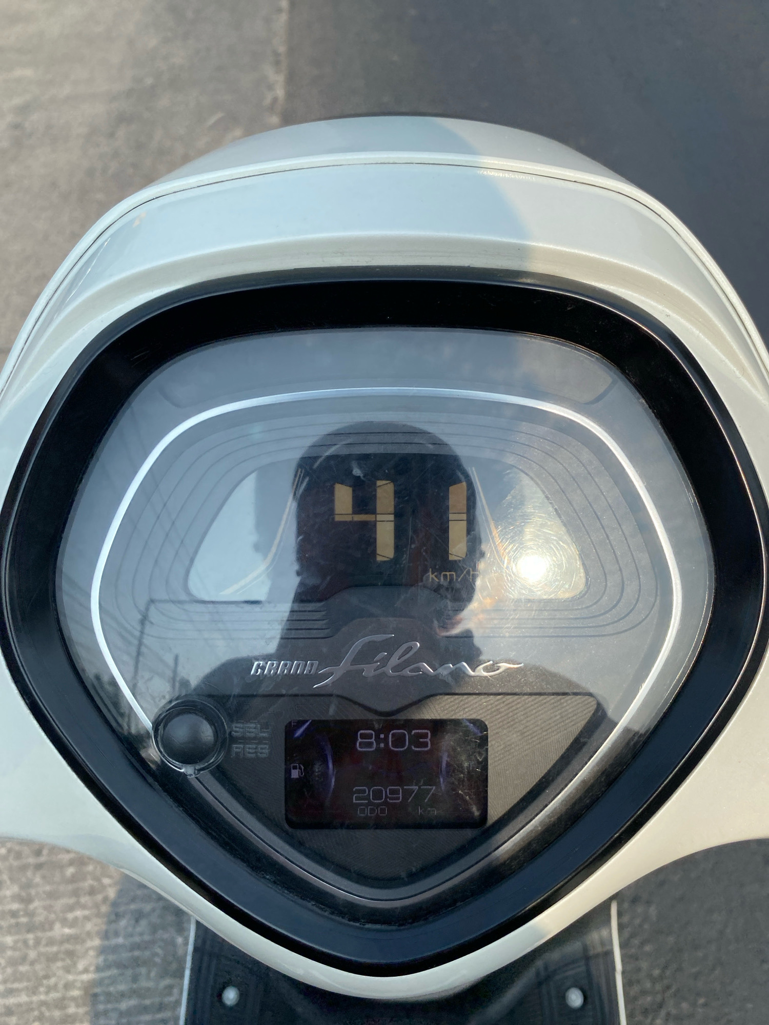 A close-up, eye-level shot captures the digital dashboard of a white scooter parked on an asphalt surface under sunlight. The dashboard displays the speed as 41 km/h, the time as 8:03, and the odometer reading as 20977 km. The brand name "GRAND Filano" is visible on the dashboard. Reflections on the screen show the rider's face and the surrounding environment. The scooter's handlebars are visible at the bottom of the image. There is no visible indication of the city of Klurak, Indonesia.