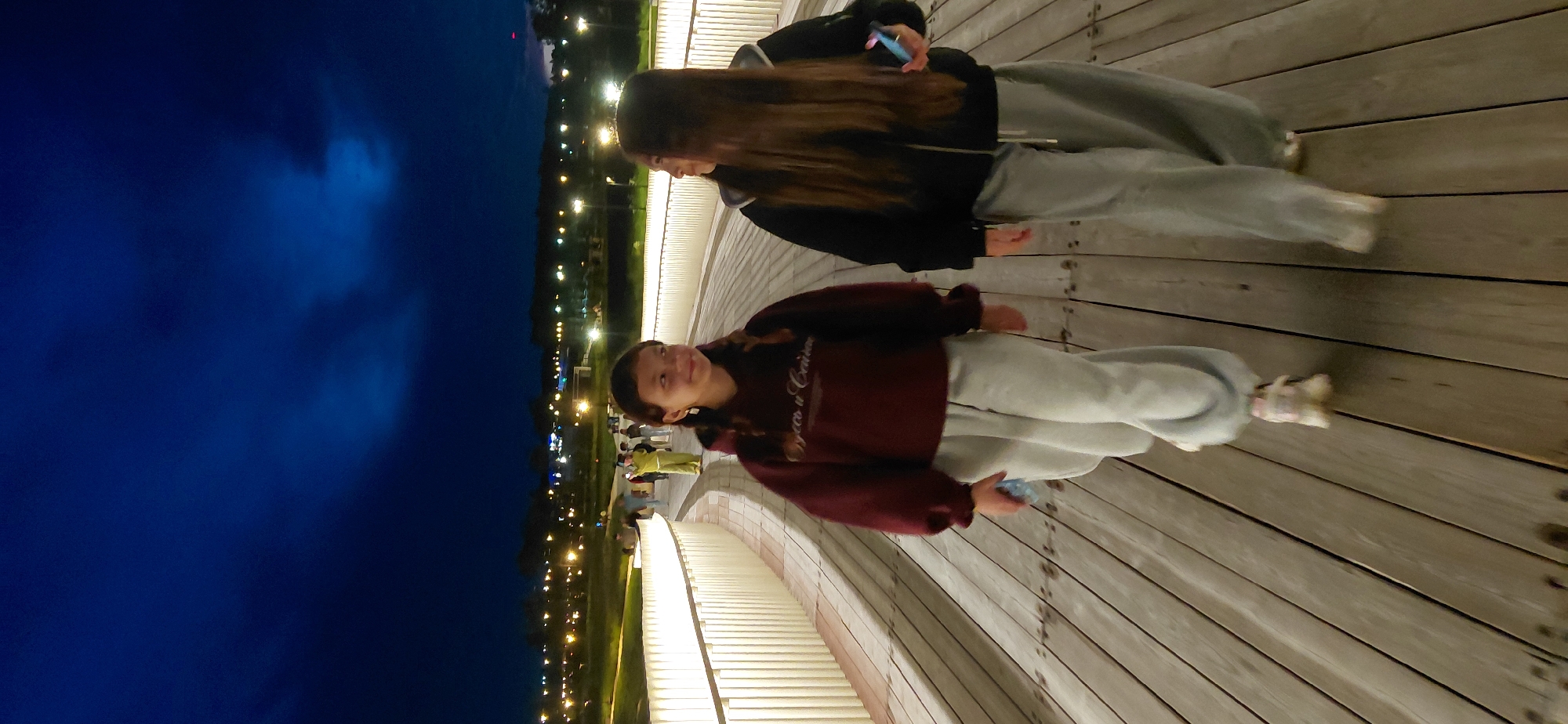 Two young females are walking along a wooden boardwalk at night. The boardwalk is illuminated by white lights lining its railing. In the background, the city of Magnitogorsk, Russia, is visible with its lights twinkling. The sky above is a deep blue, indicating it is nighttime. The female on the left is wearing a maroon hoodie and light gray sweatpants, and her hair is in two braids. She is holding a light blue object in her left hand. The female on the right is wearing a black zip-up hoodie and light gray sweatpants. Her hair is long and brown. Both are looking forward, walking away from the viewer.