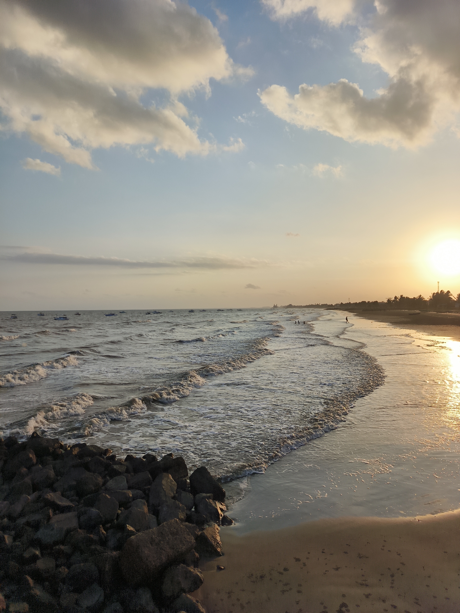 The image captures a serene sunset scene at a beach in Kupang Berkah Jaya, Indonesia. The sky is adorned with a blend of blue and warm golden hues, with scattered clouds reflecting the setting sun's light. The ocean stretches towards the horizon, with gentle waves lapping at the shore. Several boats are visible in the distance, dotting the calm sea. The beach itself is a sandy expanse, with footprints marking the wet sand near the water's edge. In the foreground, a cluster of dark, rugged rocks forms a natural barrier along the coastline. A few individuals can be seen scattered along the beach and in the shallow water, enjoying the evening. The overall atmosphere is one of tranquility and natural beauty.