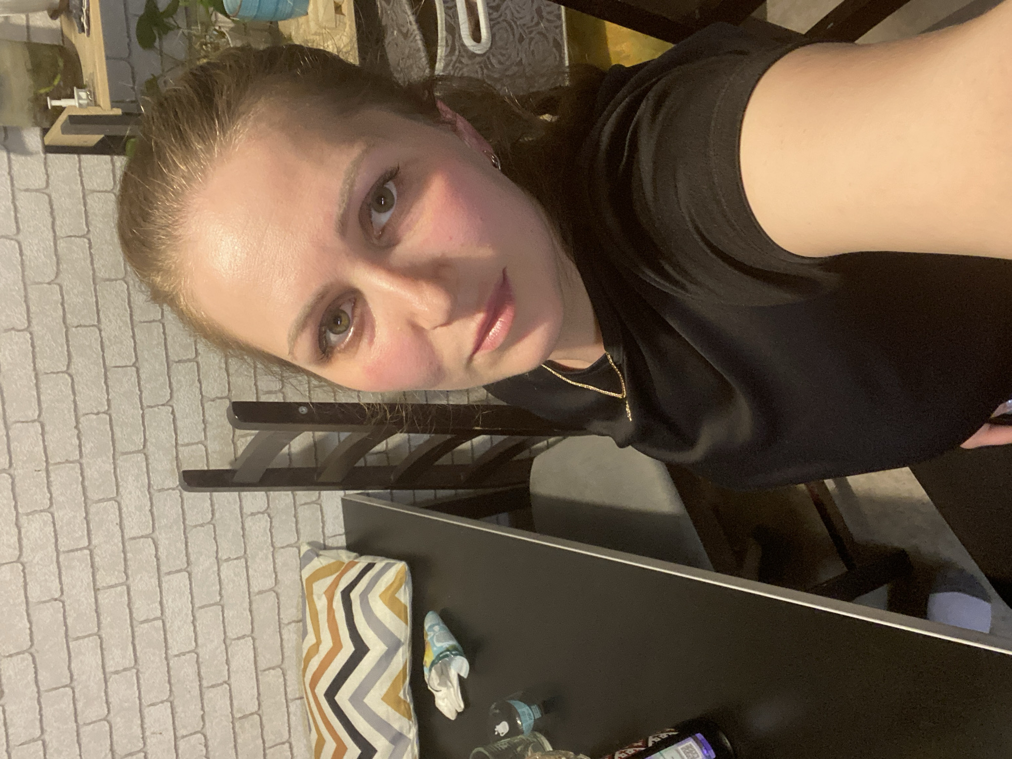 A selfie-style portrait captures a young woman looking directly at the camera. She has light brown hair pulled back, green or hazel eyes, and appears to be wearing light makeup, including blush, with rosy cheeks and light pink lips. She is wearing a black t-shirt and a delicate gold chain necklace. Her expression is calm and neutral, with a slight hint of a smile.
The image is taken indoors, with a white brick-patterned wall visible on the left side (when the image is rotated upright). Above the wall, some green plants are visible, suggesting indoor greenery. To her right, a dark table or desk occupies the lower half of the frame. On the table, there's a patterned pillow with white, grey, orange, and yellow chevron or zigzag stripes. Next to the pillow are several small items, including a crumpled tissue, a small blue and white container, and a couple of bottles, one of which appears to be a clear bottle with a dark liquid. The background behind the table is dark, showing parts of a chair or furniture legs. The lighting appears to be artificial indoor light. The location is Марковский, Russia.