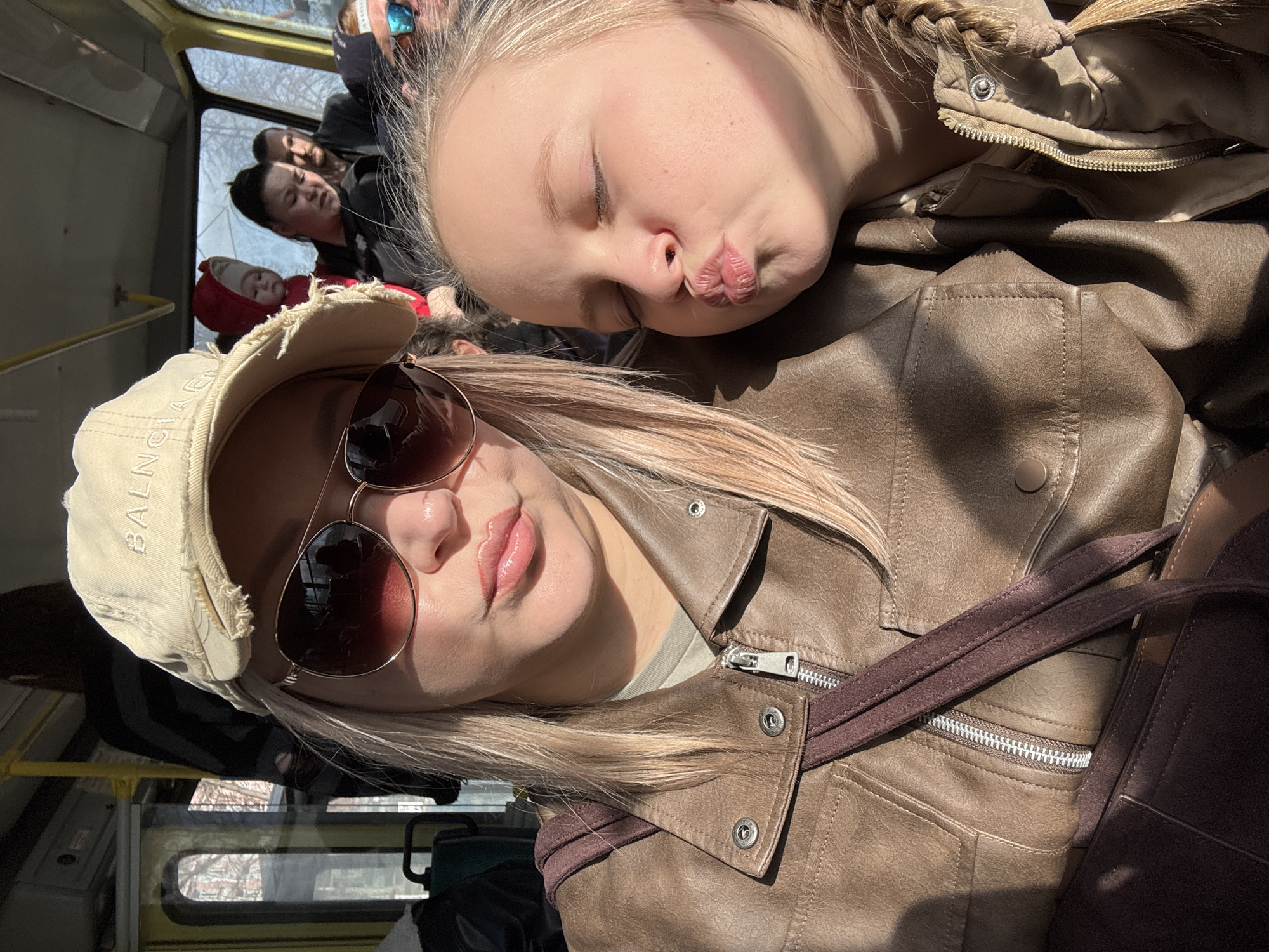 A selfie shows a woman wearing a baseball cap and aviator sunglasses next to a child, both with blonde hair, inside what appears to be a public transportation vehicle, possibly a bus or trolley. Other passengers are visible in the background. Based on the yellow railings and interior, this is likely a public transportation vehicle in Izhevsk, Russia. The woman and child are wearing brown leather jackets. The child is pouting their lips and has closed eyes. The woman is gazing at the camera.