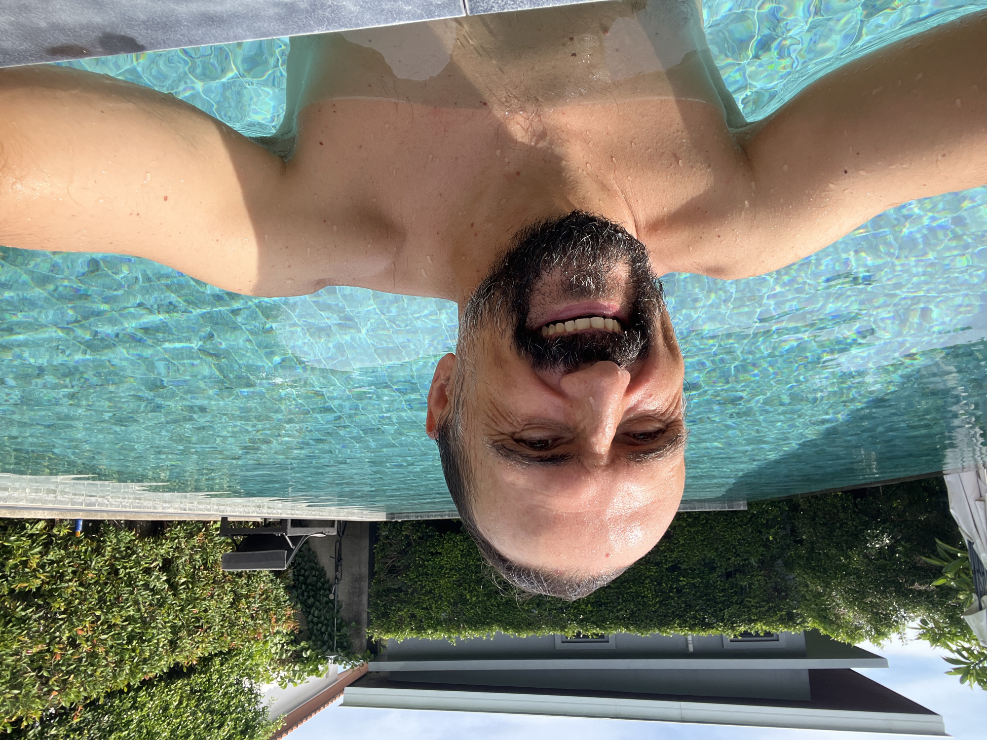 A man with a beard is swimming in a pool, smiling at the camera. The pool is surrounded by greenery and a building. The photo was taken in บ้านบางรักษ์, Thailand.