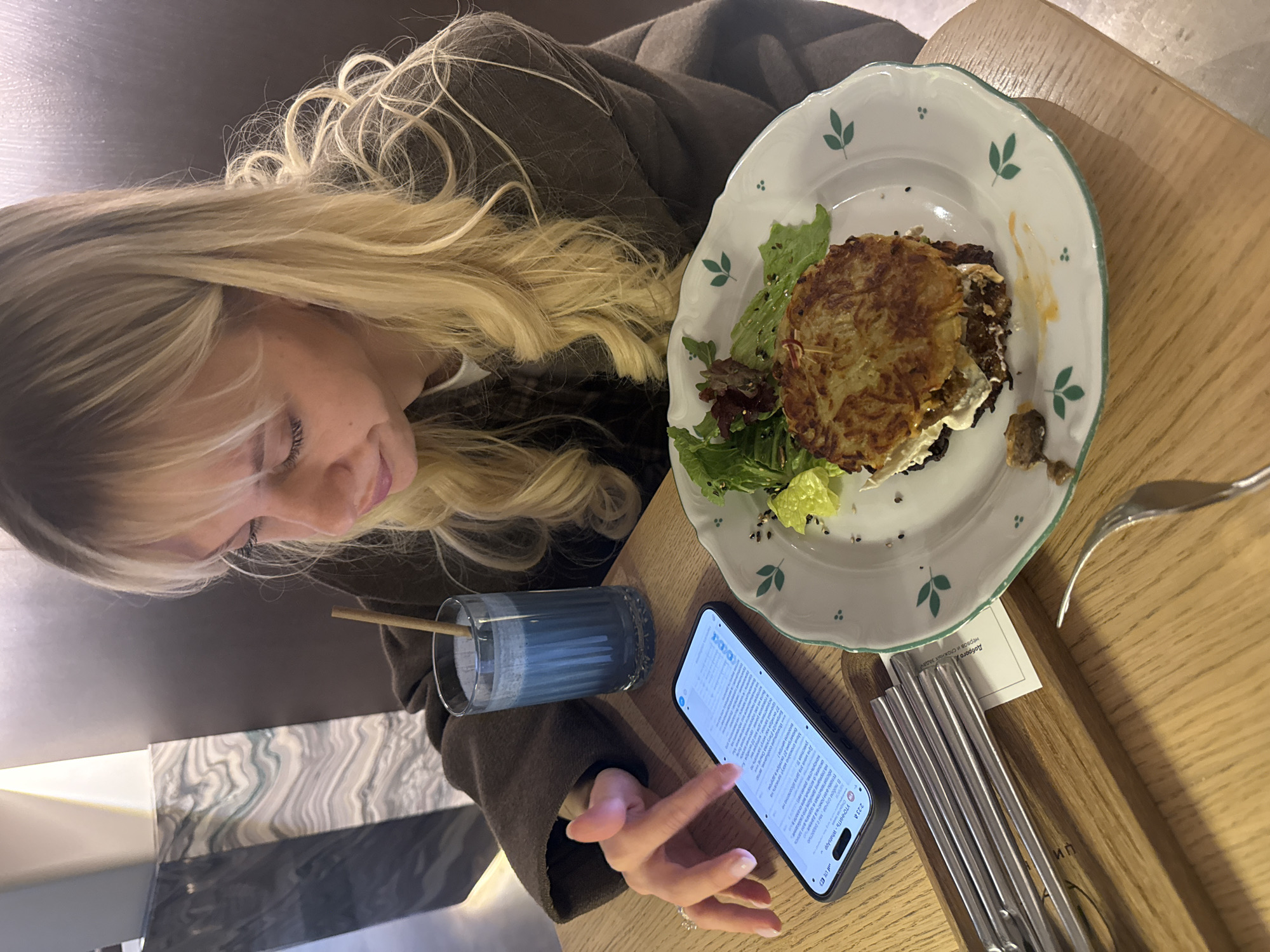 A woman with long blonde hair is seated at a wooden table. She is looking at her phone, which is displaying a WhatsApp chat in Russian. Next to the phone is a glass of blue liquid with a straw. A plate with a potato pancake, salad, and some other toppings is also on the table, along with silverware. The text on the phone mentions "Moscow" and "Russia," indicating the location.