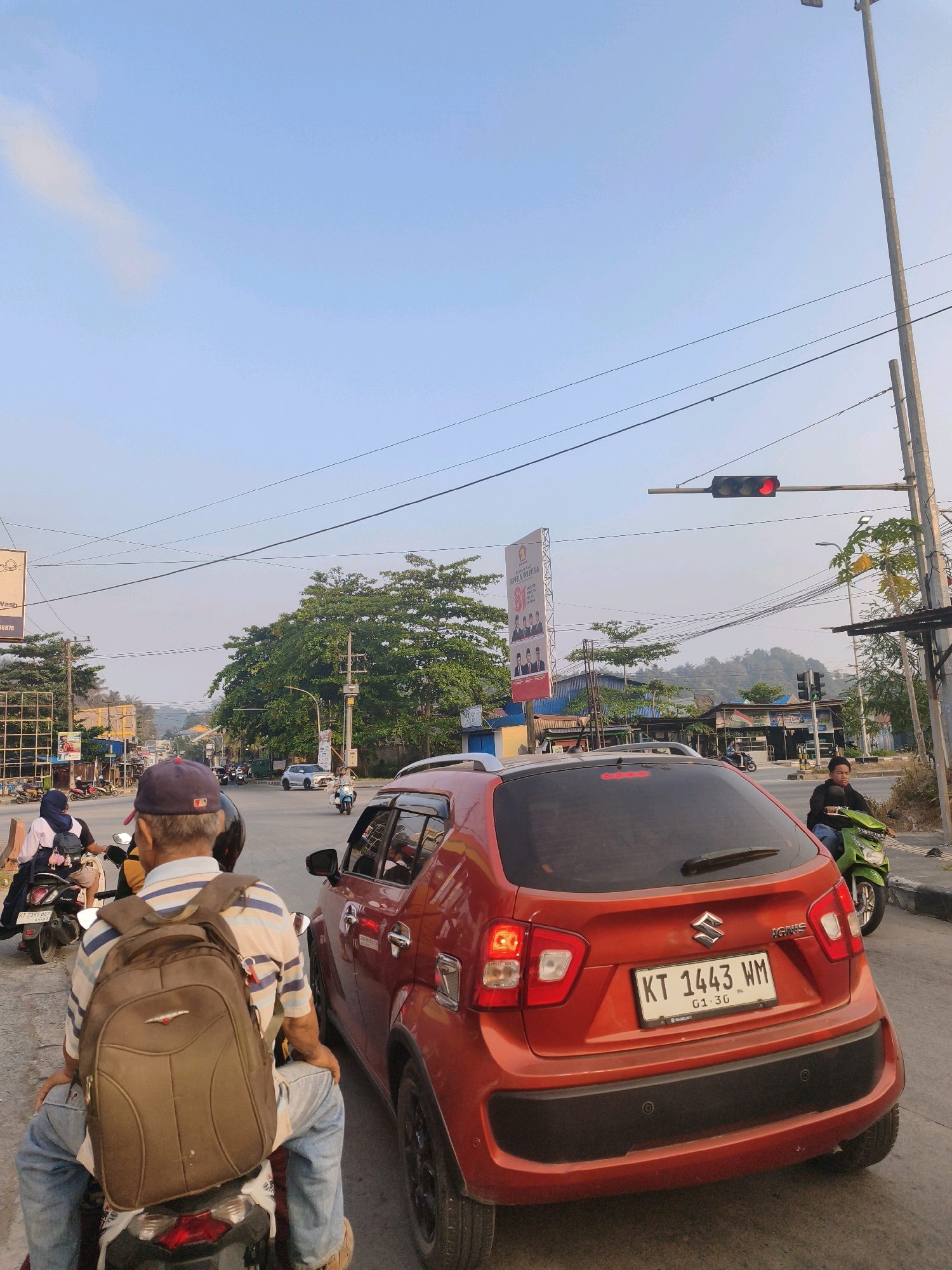 The image depicts a traffic intersection in Samarinda, Indonesia. In the foreground, a man wearing a blue and white striped shirt, blue jeans, and a backpack is seated on a motorcycle. Beside him, to his right, is a red Suzuki Ignis car. The car's license plate reads "KT 1443 WM 01-30". In the background, other motorcycles and cars are visible, suggesting a moderate traffic flow. A traffic light displaying a red signal is visible above the intersection. Several buildings and lush green trees line the street, indicating an urban environment. A large banner with text and images of people is also present, likely for a political or promotional event. The sky is clear and blue with a few wispy clouds. The overall scene captures a moment in the daily life of Samarinda, characterized by its busy streets and tropical vegetation.