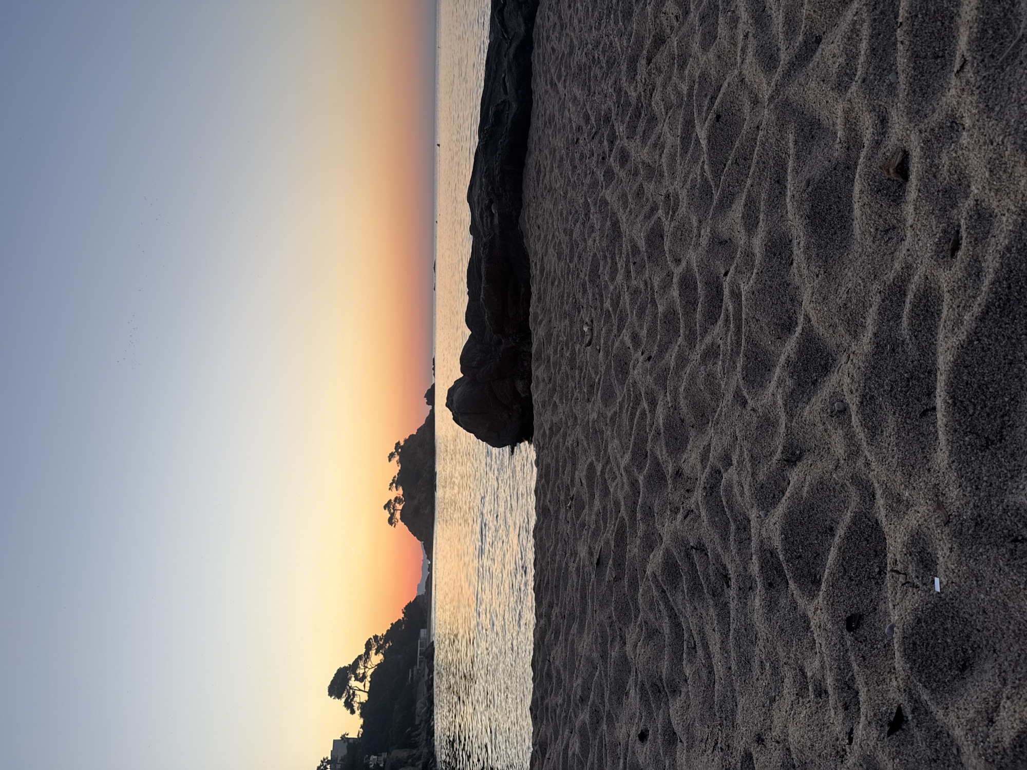 The image depicts a serene beach scene at sunrise in Castell d'Aro, Platja d'Aro i s'Agaró, Spain. The foreground is dominated by textured sand dunes, showing the ripples and patterns left by the tide. A large, dark rock formation sits in the middle ground, partially obscuring the calm sea. In the distance, a small island with several trees is visible, silhouetted against the gradient sky. The sky transitions from a pale blue at the top to warm hues of orange and yellow near the horizon, indicating the early morning sun. The sea reflects the colors of the sky, creating a tranquil atmosphere. The coastline to the left features vegetation and structures, suggesting a developed area beyond the natural beach. The overall scene is peaceful and devoid of people.