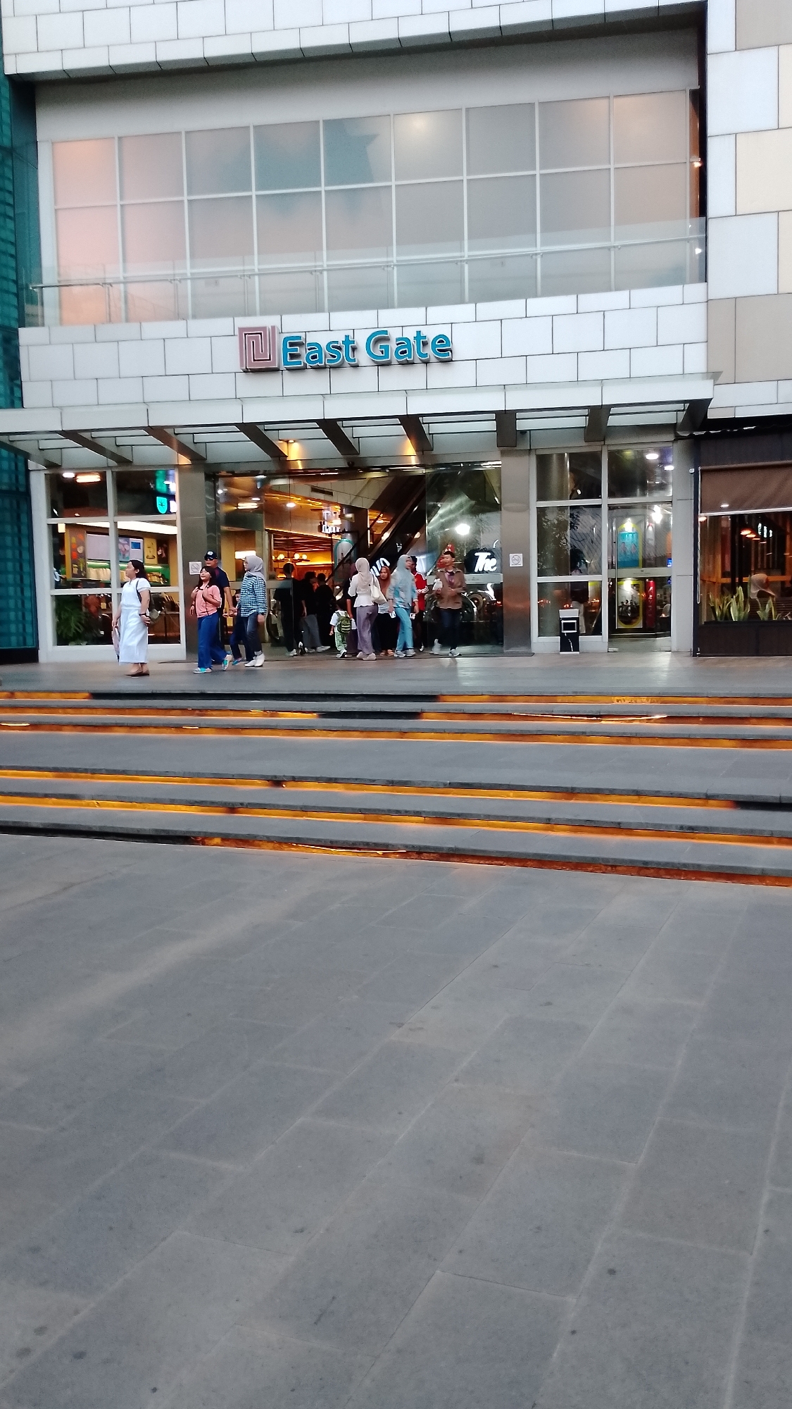 A street-level, medium shot captures the entrance to a modern building labeled "East Gate" in South Tangerang, Indonesia. The facade is characterized by a white tiled base and a glass-enclosed upper level. A prominent sign with a stylized "E" logo and the words "East Gate" in blue text is centered above the entrance. Below the sign, a covered walkway leads into the building's interior, which is visible through large glass doors and windows.
A group of people, diverse in age and attire, are gathered near the entrance, some appearing to be queuing. A woman in a white dress stands to the left, facing away from the camera. Others are dressed in casual clothing, including shirts, pants, and headscarves. The foreground features a wide, tiered pathway with illuminated orange steps, leading up to the building's entrance. The overall impression is a bustling urban scene outside a commercial establishment.