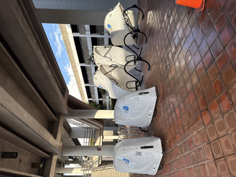 The image presents an outdoor scene located in Hialeah, United States, specifically beneath an elevated concrete transit structure. The ground is surfaced with reddish-brown square tiles, exhibiting wet areas that reflect ambient light. Five individual bicycle storage units, manufactured by PearDeck and displaying a white speckled finish, are arranged on the tiled pavement. Two units on the left are in a closed position, while the three units to their right are open, revealing internal black metal frames designed to secure bicycles. Behind the closed units, sections of conventional silver U-shaped bicycle racks are partially visible. An orange traffic cone rests on the ground in the lower right foreground. A small, faint white heart shape is drawn on a wet tile near the closed bicycle pods. In the midground, concrete support columns and staircases with metal railings ascend, connecting to the elevated structure overhead. The background features a multi-story concrete building, consistent with a parking garage, visible beneath a partly cloudy blue sky. No individuals are present in the depicted scene.