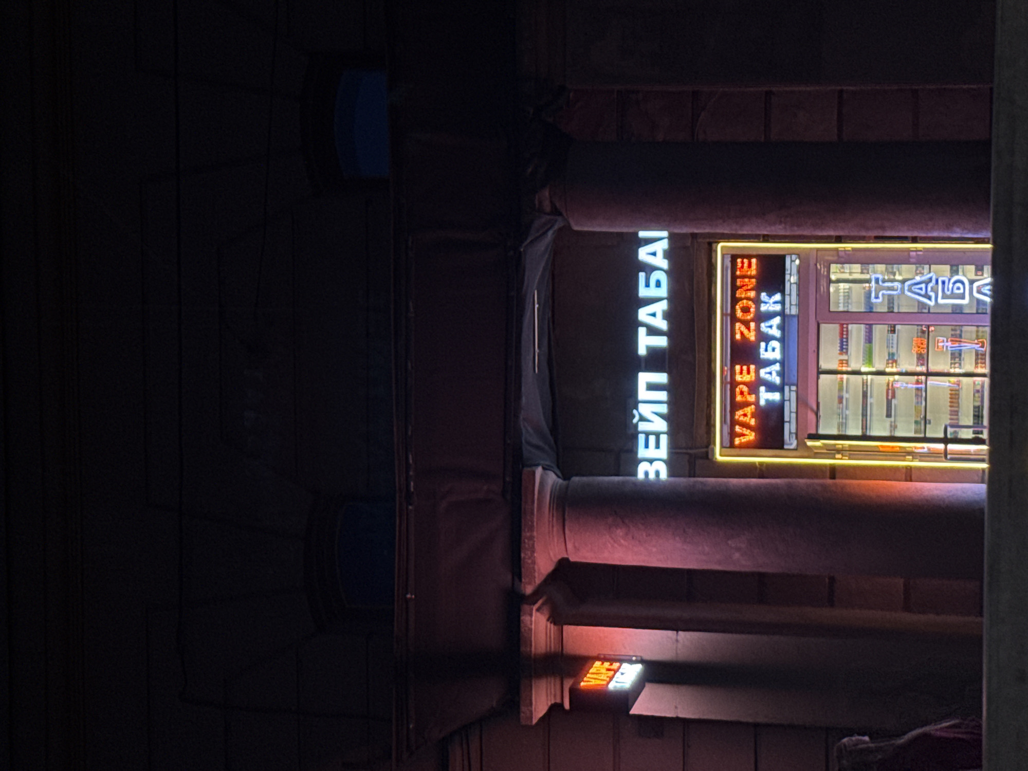 The image shows a neon sign that reads "VAPE ZONE ТАБАК" in Russian, illuminated at night in front of a store. The store appears to be a tobacconist or vape shop. The sign is mounted above the entrance to the store, which is framed by a yellow neon light. Inside the store, shelves stocked with products are visible behind a glass door. The store is flanked by two large columns. The overall scene suggests an urban setting, likely in a city with Cyrillic signage, such as Saint Petersburg, Russia, given the presence of the Russian text. A person is partially visible on the left side of the image, dressed in dark clothing and a red garment. The background is dark, indicating it is nighttime.