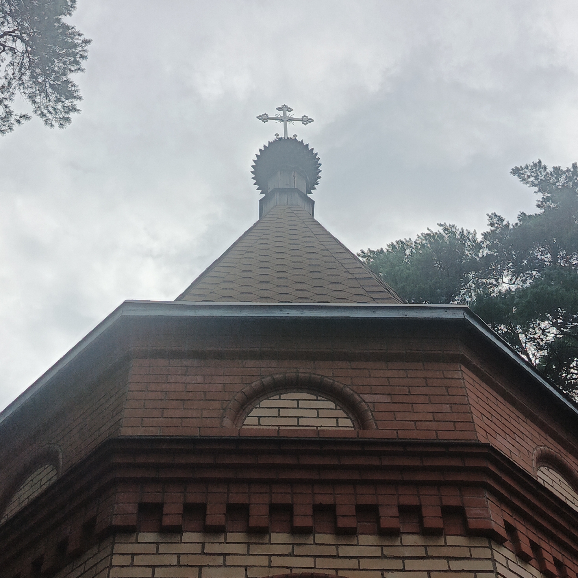 A low-angle, close-up, outdoor photograph shows the top portion of a brick building, likely a chapel or small church, against an overcast sky. The building has a steeply pitched, shingled roof. The brickwork features alternating courses of light and dark red bricks, with decorative brickwork patterns near the eaves. A prominent arched window with a light-colored brick infill is visible on the building's facade. At the apex of the roof, a dark, multi-tiered spire is topped with an ornate, silver-colored Christian cross. Pine trees are visible in the background, some obscuring the sky with their branches, suggesting the building is located in a wooded area. The lighting is diffused due to the cloudy conditions. This structure is identified as the chapel of St. Nicholas in Nikolo-Beryozovka, Russia.