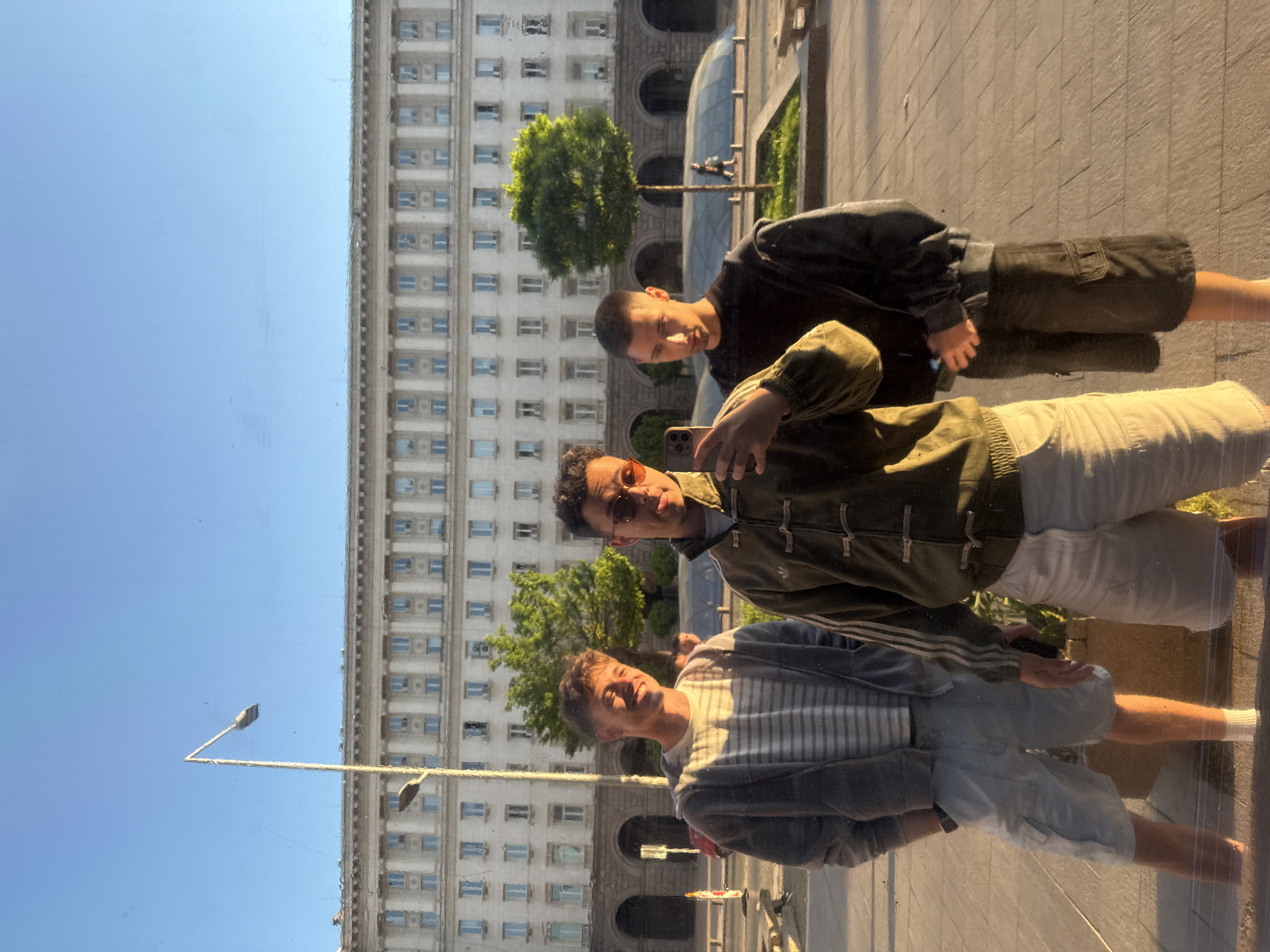 Three young men are reflected in a glass surface against a backdrop of a large, pale building. The building features rows of windows and arched entryways. A tree is visible near the center of the building. The sky above the building is clear and blue. The men are positioned closely together. The man closest to the viewer is wearing a striped shirt and light-colored shorts, covered by a jacket. The second man is wearing sunglasses, an olive-green jacket, and white shorts, holding a phone. The third man is wearing a dark shirt and dark shorts. The location is likely Sofia, Bulgaria, based on the architectural style of the building, similar to the Largo complex.