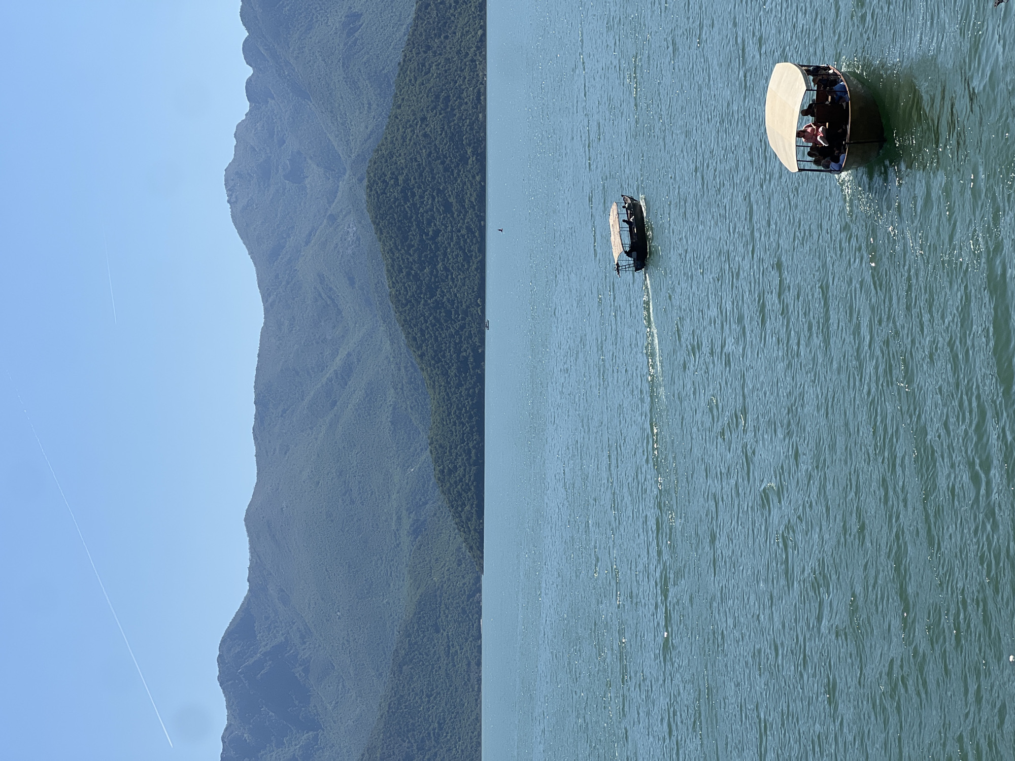 The image shows two boats on a lake near Kruševica, Montenegro, with mountains in the background. One boat is larger, with a white canopy and several people onboard, while the other is smaller and appears to be moving faster, creating a wake. A jet trail is visible in the blue sky. The water is slightly choppy.
