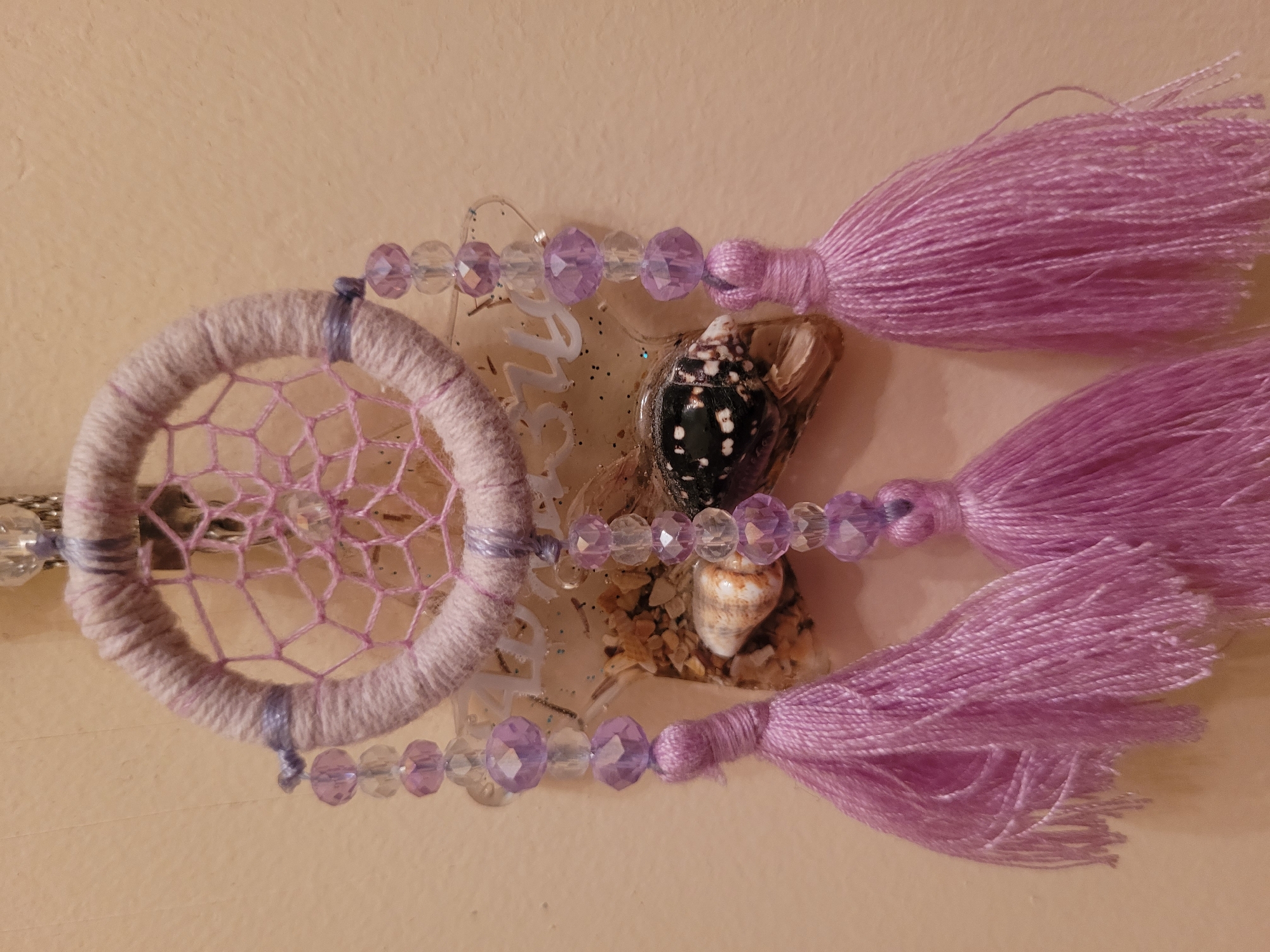 A close-up shot presents a handcrafted dreamcatcher against a pale beige background. The dreamcatcher features a circular frame wrapped in light purple yarn, with a loosely woven web of similar yarn stretched across the center. Dangling from the frame are strings of clear and light purple faceted beads, interspersed with small seashells and three large tassels of light purple thread. The seashells and beads appear to be attached to a transparent acrylic or resin base. The overall color scheme is dominated by shades of purple and beige. The scene does not show any elements that allow identifying the location of Izhevsk, Russia.