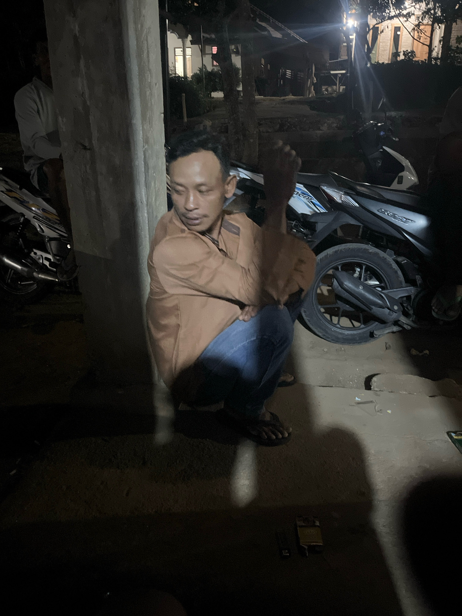 An adult male is crouching on the ground next to a concrete pillar at night. He is wearing a light brown collared shirt and blue jeans. He is looking downwards and to his right. Behind him, to his right, is a dark-colored motorcycle. To his left, further back, is another motorcycle, this one white and black. In the background, there are dimly lit structures, possibly houses, with lights visible. The ground appears to be dirt or gravel. The image seems to have been taken outdoors in a residential or semi-rural area. The location is identified as Kebumen, Indonesia.