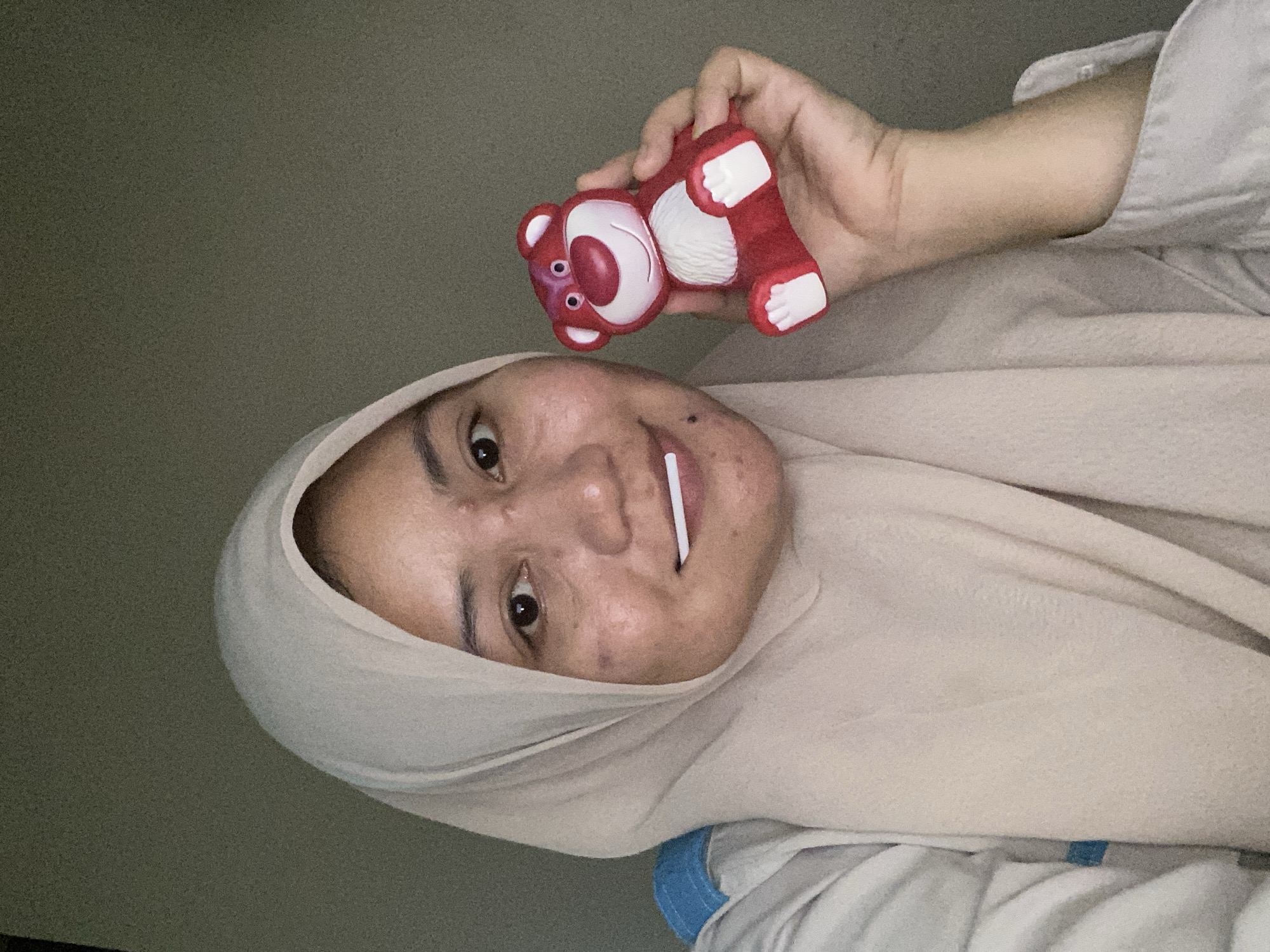 The media file is a selfie featuring a person wearing a light-colored hijab. The person's face is partially visible, and they are holding a small, red toy bear with white accents in their right hand. The toy appears to be Lotso Huggin' Bear from Toy Story. The person has a white object, possibly a straw or a piece of candy, in their mouth. Their expression is difficult to discern due to the image orientation and quality, but they appear to be looking at the camera. The background is a plain, dark wall. The lighting suggests it might be indoors, possibly in the evening or with artificial light. There is no visible text in the image. The setting appears to be casual and personal, given the nature of a selfie and the toy. The location context is Banjarbaru, Indonesia, but there are no specific visual cues within the image to confirm this.