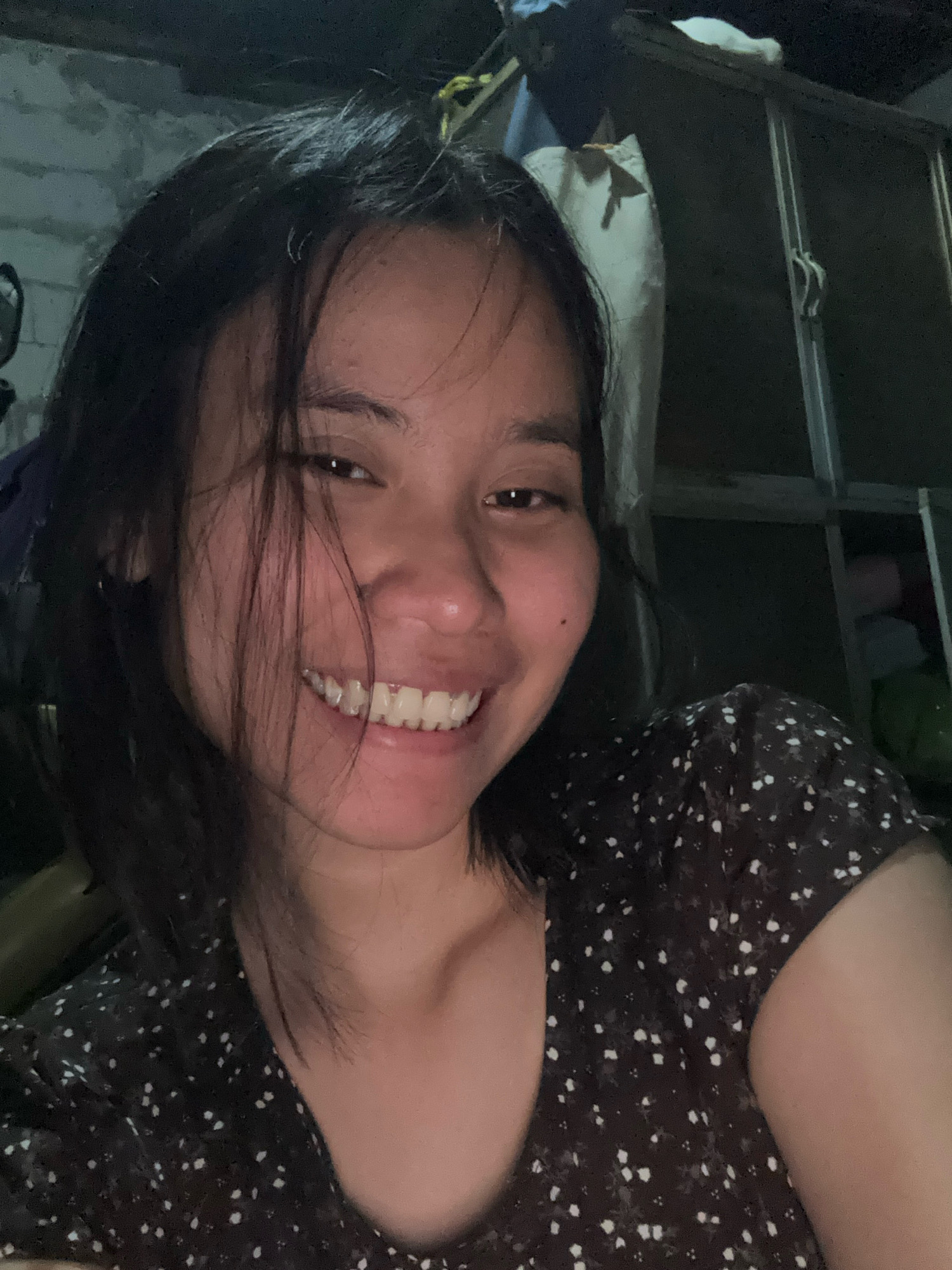 A young woman is captured in a close-up, selfie-style photograph. She has dark hair that is slightly disheveled and frames her face. Her eyes are dark, and she is smiling broadly, revealing her teeth. She is wearing a dark-colored top with a white floral pattern. The background is dimly lit and appears to be an indoor setting, possibly a room with some clutter visible, including what looks like construction materials or stored items. The overall impression is casual and personal. This image was taken in East Jakarta, Indonesia.