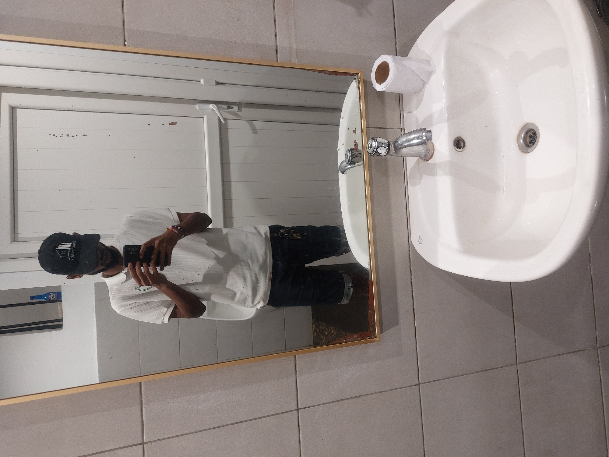 The image shows a bathroom scene, captured through a mirror reflection. A person wearing a baseball cap, a white polo shirt, and dark jeans is holding a phone, presumably taking the photo. The background reflected in the mirror shows a white door with horizontal panels and a bottle on a shelf. A white sink is positioned beside the mirror, complete with a faucet and a roll of toilet paper mounted above it. The bathroom floor and walls are tiled with gray squares. The absence of specific location markers or architectural styles does not allow for confirmation of the location in Dar es Salaam, Tanzania, nor can the specific date be identified.