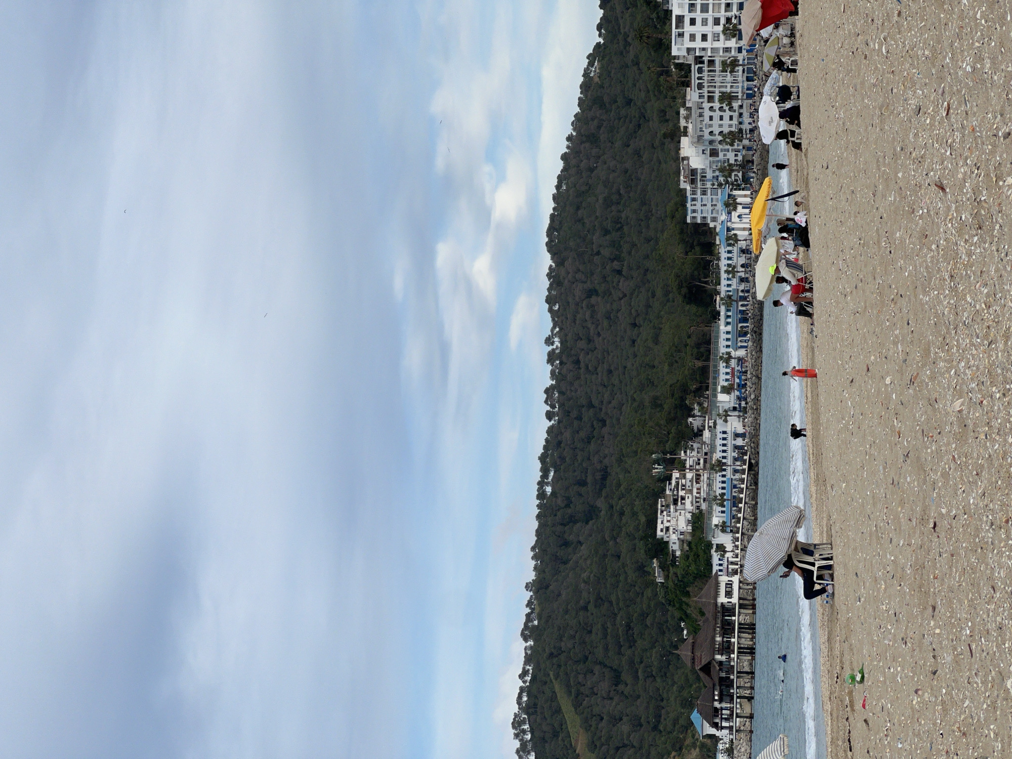 The image shows a beach scene in M'Diq, Morocco. The beach has a sandy surface with scattered seashells. Several people are on the beach, some under umbrellas and some in the water. Buildings are visible near the beach. A forested hill forms a backdrop. The sky is partially cloudy.