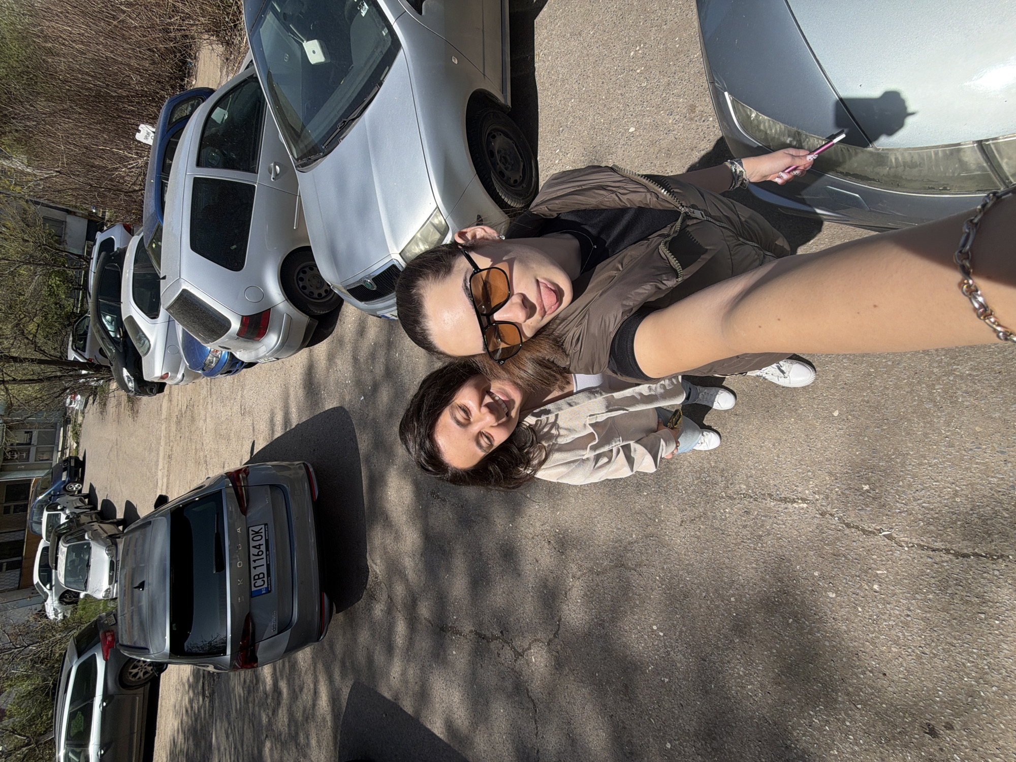 The image captures a rotated outdoor selfie of two women in Sofia, Bulgaria. The woman in the foreground, taking the picture, wears a brown puffer vest over a dark top, dark-rimmed sunglasses with amber lenses, and has her hair pulled back. She is playfully sticking her tongue out. Her left arm, adorned with a silver chain bracelet, is visible extending towards the camera. Her right hand, with manicured nails, holds a pink pen or brush near the headlight of a silver car.
Positioned slightly behind and below her is the second woman, with dark hair, smiling broadly with sun-squinted eyes, wearing a light beige jacket.
The background shows a street or parking area with asphalt pavement and numerous parked cars. Distinct shadows on the ground indicate bright, clear daytime weather. Visible in the background are trees and shrubs with sparse foliage, and a building in the distance. One prominent gray Skoda car displays the license plate "CB 1164 OK," with "SKODA" written on its rear, confirming the "CB" Sofia registration. The overall mood is casual and cheerful.