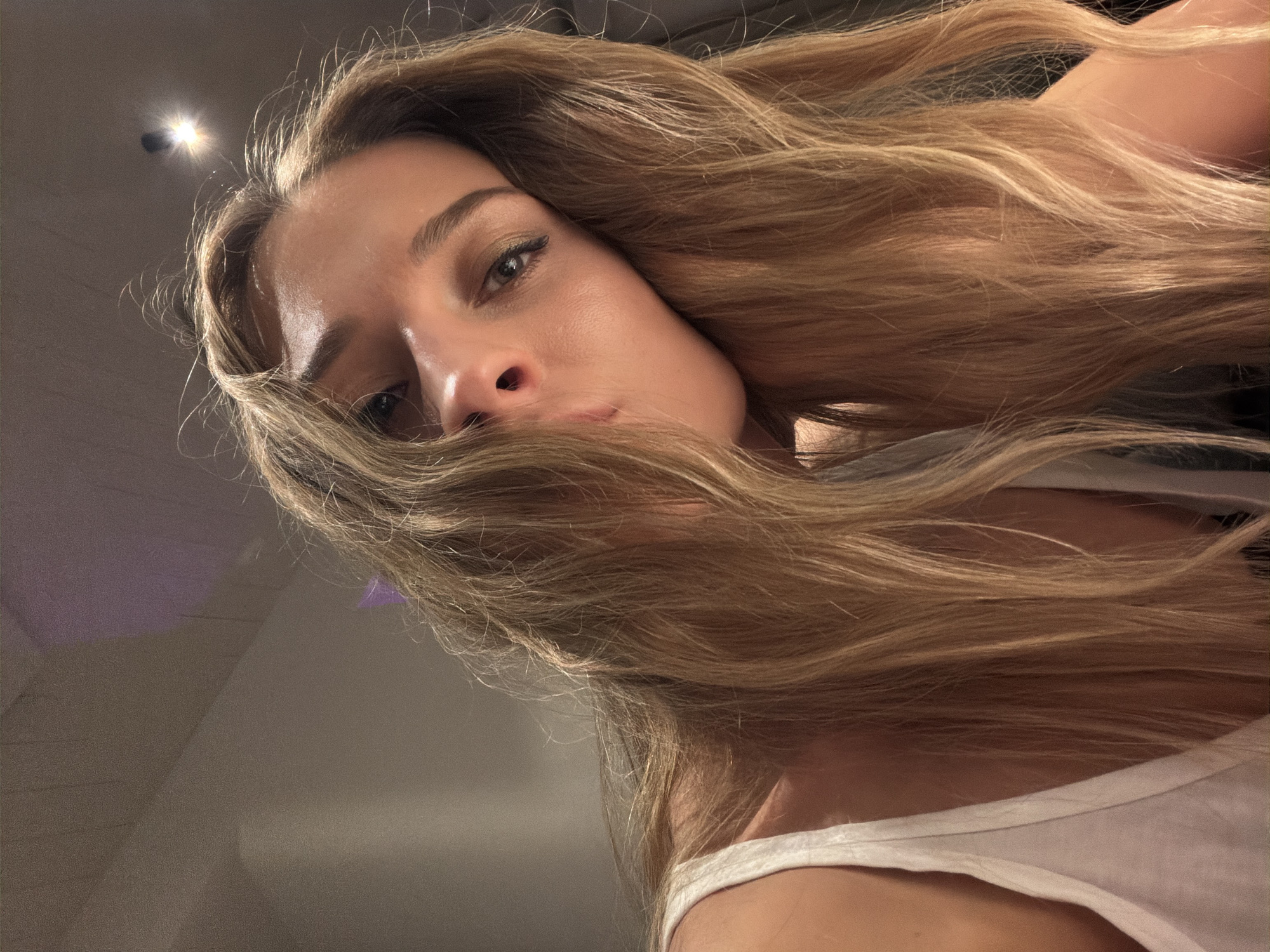 The image shows a woman with long, blonde hair partially covering her face. She is wearing a white tank top. The background is blurry but appears to be an indoor setting. There is no indication of the location being Moscow, Russia or any other specific city.