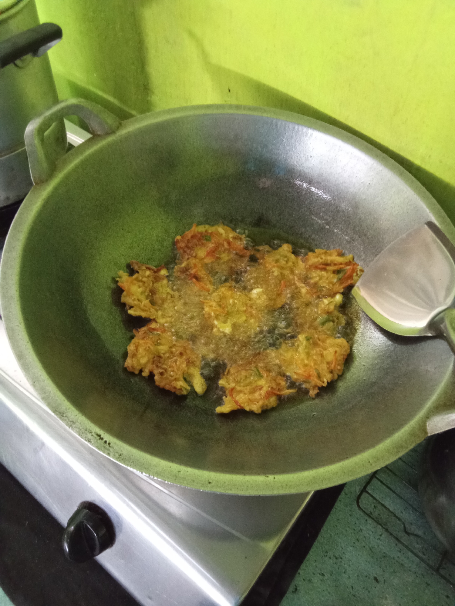A close-up, eye-level shot captures fritters frying in a wok, which sits on a gas stove. The wok, made of grey metal, is filled with bubbling oil and several golden-brown fritters. A silver spatula with a green handle rests on the right side of the wok. The background is a bright green wall. The image appears to have been taken in a kitchen, likely in Subang, Indonesia, as suggested by the presence of a common cooking setup and the overall ambiance.