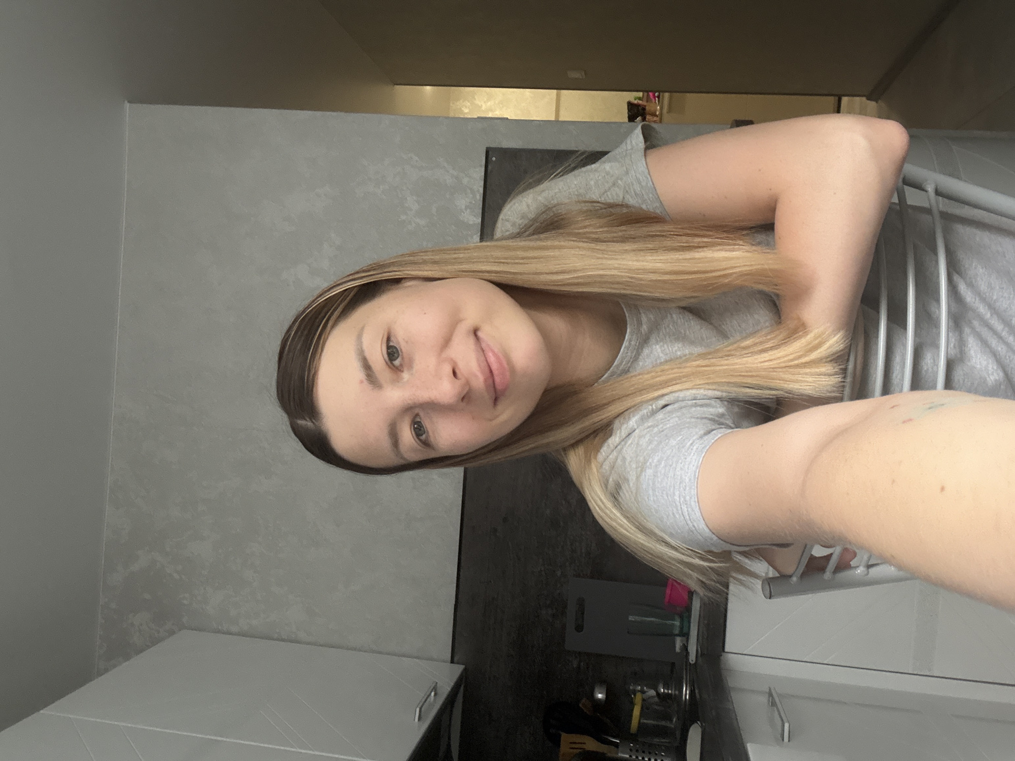 A young woman with blonde hair, wearing a grey t-shirt, is sitting on a chair in a kitchen. Her arms are crossed in front of her and resting on the back of the chair. The kitchen has white cabinets and grey textured walls. There is a sink and countertop visible with various kitchen items. The lighting suggests it is daytime. The overall scene is domestic and casual. The image location in Moscow, Russia, is not determinable from the visual information provided.