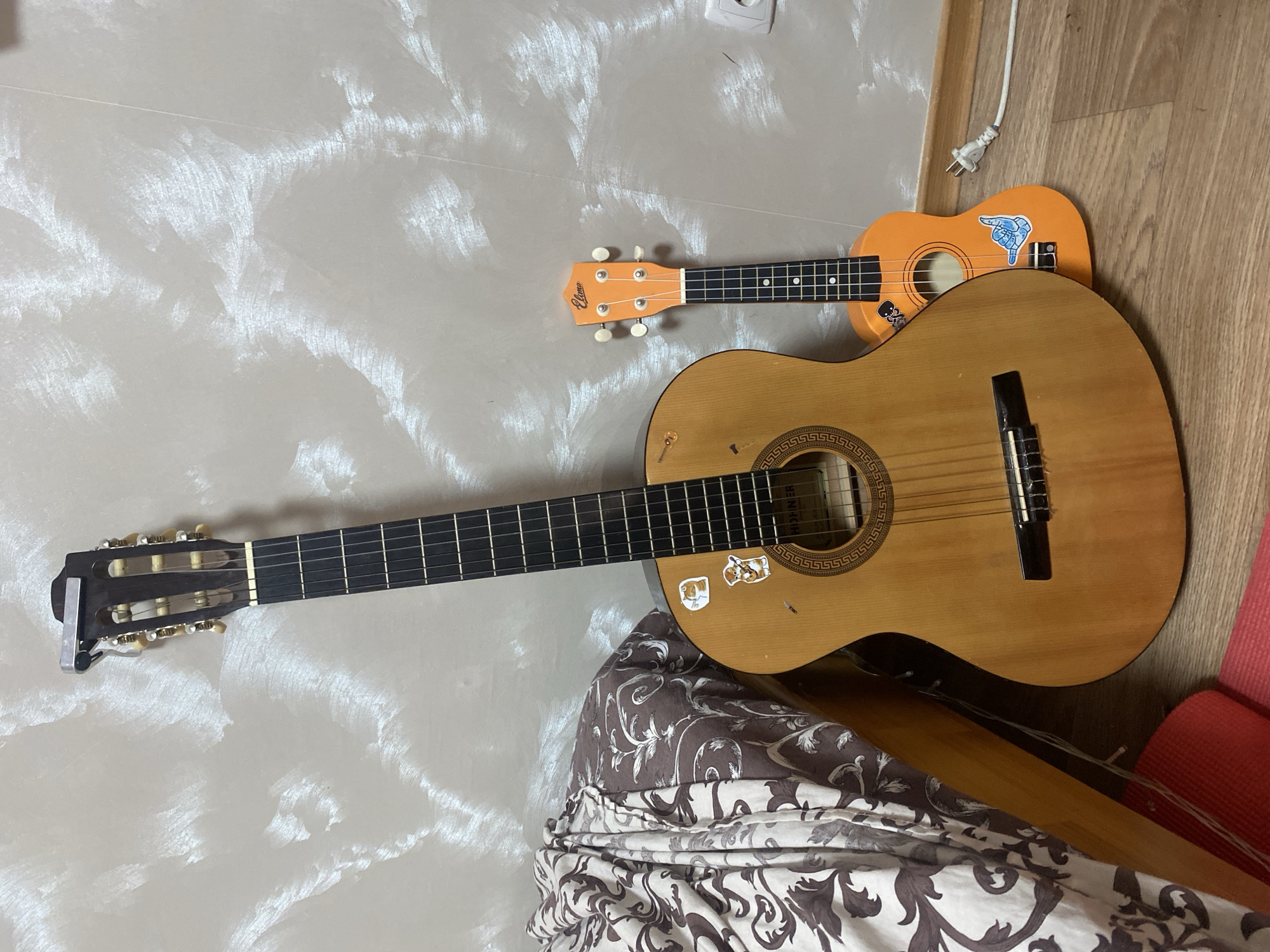 In Chornomorsk, Ukraine, two string instruments, a guitar and a ukulele, stand against a textured wall. The larger instrument, a classical guitar with a natural wood finish, is positioned to the left and is slightly angled. It has a dark headstock with tuning pegs and a capo attached. Several stickers are visible on its body, including what appears to be a small cartoon cat and a circular emblem. To the right of the guitar is a smaller, bright orange ukulele. The ukulele has a white headstock with tuning pegs and a label that reads "Elina". It also features stickers, one of which is a blue hand gesture and another a butterfly. A white electrical cord with a plug runs along the baseboard on the right side of the image, near the ukulele. A portion of a bed with patterned bedding is visible on the left, behind the guitar. A red rolled-up mat lies on the floor in the foreground. The wall has a distinctive textured pattern with brushstroke-like highlights.