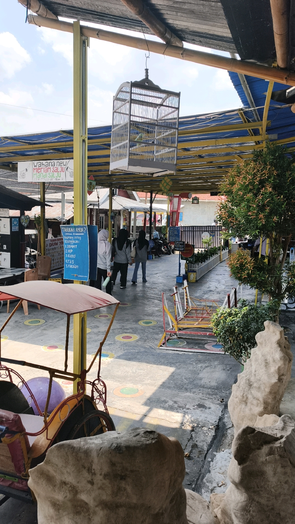 The image shows an outdoor walkway at a facility in Air Paoh, Indonesia, possibly a recreational area or market. A large birdcage hangs from the ceiling, containing at least one bird. Several people, primarily women wearing hijabs, walk along the paved pathway, which is marked with colorful circles. A traditional Indonesian pedicab is parked on the left side of the frame, and a play structure with colorful bars is visible further down the walkway. The area is covered by a roof supported by yellow and blue beams, with natural lighting filtering through. Lush green foliage and decorative rocks are also present, adding to the environment. Signs in Indonesian are visible, one of which lists items such as "Dompet" (wallet), "HP" (mobile phone), and "Kunci" (keys).