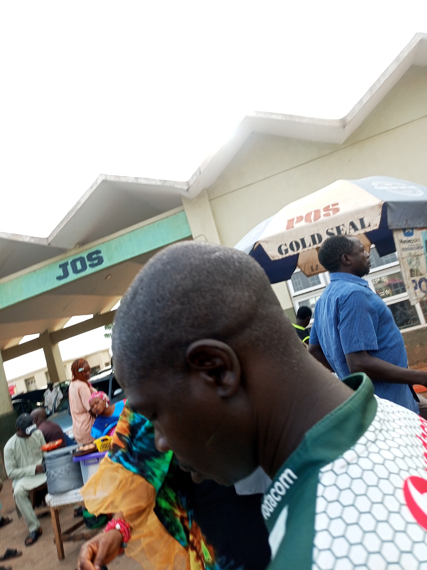 In Gombe, Nigeria, a scene unfolds under a bright, overcast sky in front of a building with a distinctive green facade bearing the blue lettering "JOS." The building has a modern, angular roofline. In the foreground, the back of a person's head is prominent, their dark hair short and textured. They are wearing a green and white patterned shirt with a honeycomb design and the word "Vodacom" visible on the collar.
To the right of the foreground figure, a man in a blue striped shirt stands facing away from the camera, looking towards the right. He is partially obscured by an umbrella advertising "POS GOLD SEAL." Behind him, a group of people are visible, including a woman in a light pink hijab and a peach-colored outfit, leaning on a dark-colored vehicle. Another person, possibly a man, is seated on a stool to the left, wearing a light green garment. In the middle ground, a table holds a few items, including what appears to be food arranged on a tray and a white plastic container. The ground surface appears to be dirt or gravel. The overall impression is of an outdoor public space, possibly a market or a transportation hub, with people engaged in various activities.