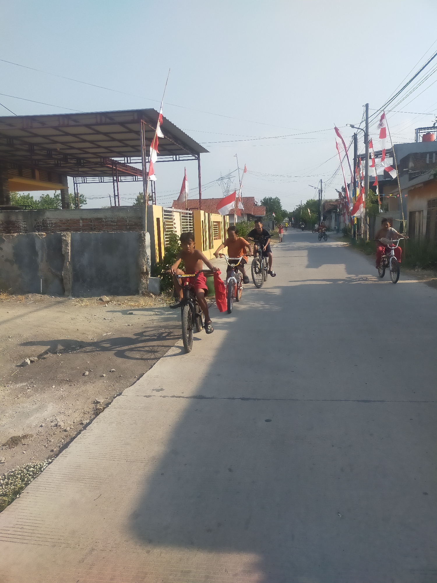 In Sumingkir, Indonesia, a group of young boys are riding bicycles down a paved street. The street is lined with houses and decorated with numerous Indonesian flags, indicating a celebration. One boy in the foreground, wearing red shorts, is looking towards the camera as he rides. Other children are further down the street, also on bicycles. The sky is clear and blue.