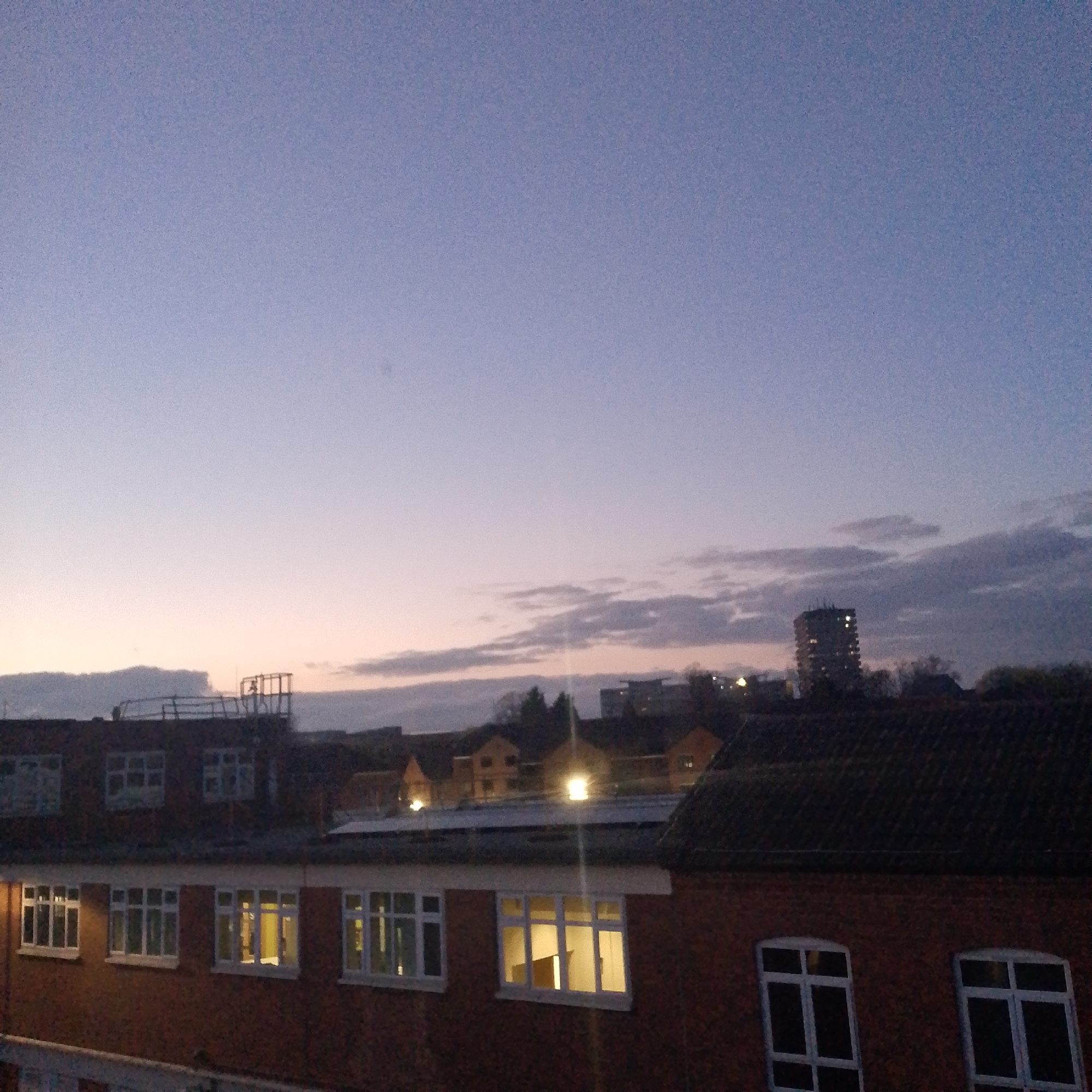 The image captures a twilight cityscape in Coventry, United Kingdom. The foreground is dominated by a red brick building, likely a multi-story structure, with numerous windows, some illuminated from within. Above this building, a skyline of various structures is visible, including a tower block with several lit windows, smaller residential buildings, and hints of trees. The sky transitions from a darker purple-blue at the top to a lighter orange-pink near the horizon, indicating either sunrise or sunset. Clouds are scattered across the sky, adding texture to the scene. The overall impression is of a quiet, urban environment during the transition from day to night or vice versa.