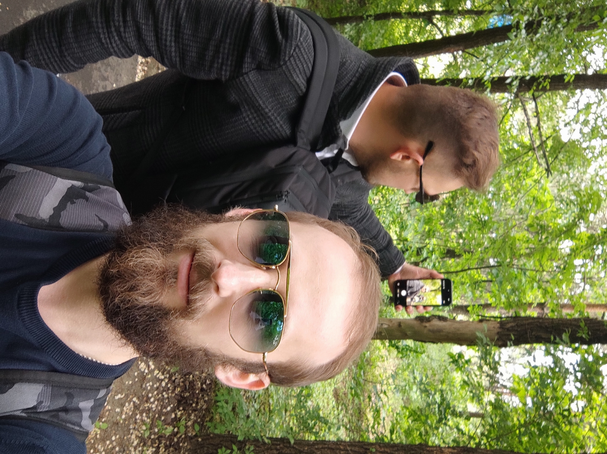 The image shows two men outdoors, in what appears to be a park or wooded area. The man in the foreground is wearing sunglasses and a dark blue shirt with a gray camouflage pattern on his backpack strap. He has a beard. The second man, slightly behind, is holding a smartphone and taking a picture. He is wearing glasses, a black jacket with a hood, and a white shirt. Trees and foliage are visible in the background. The location is likely in a park in Moscow, Russia.