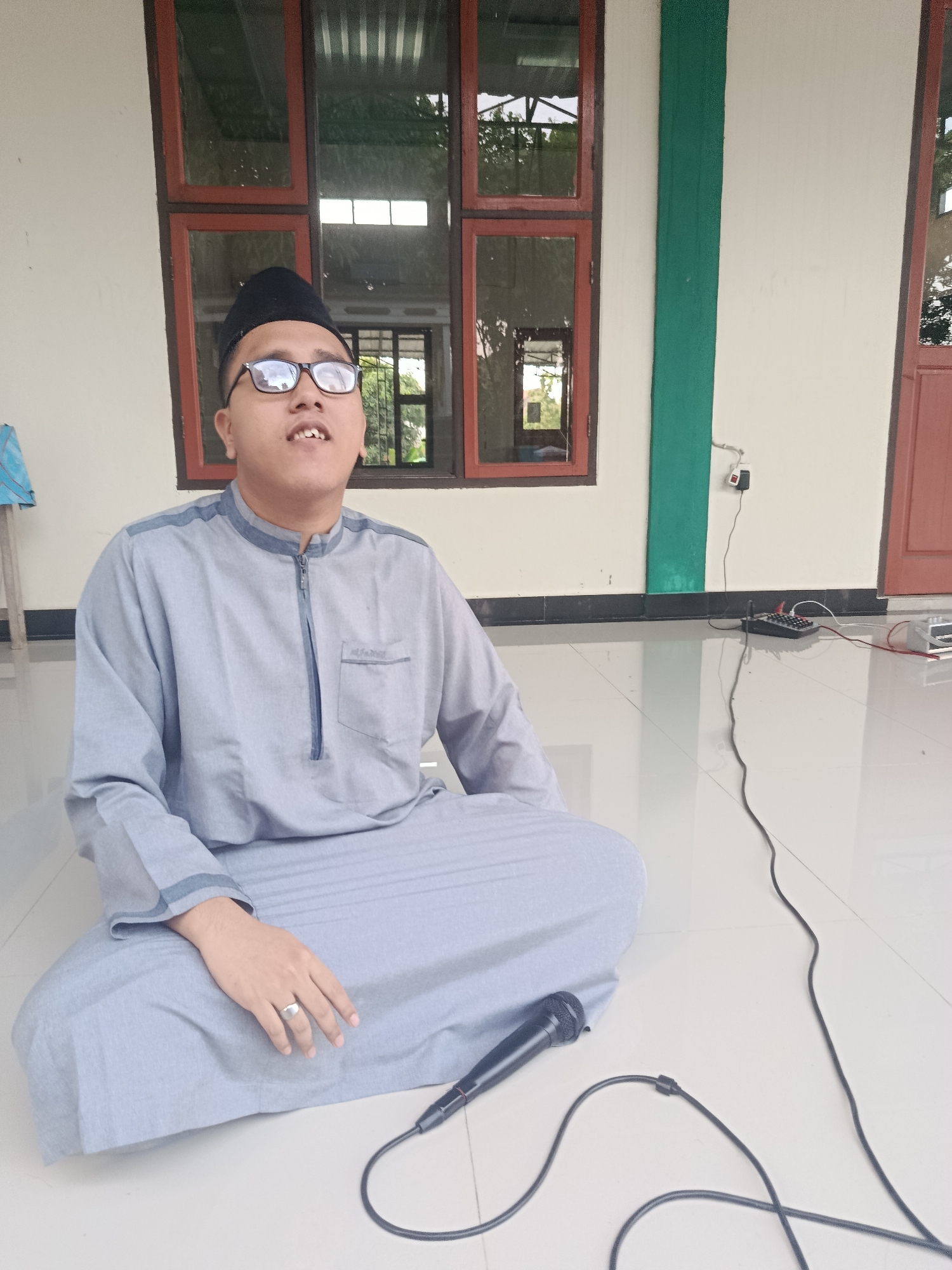A young man wearing a black skullcap, glasses, and a light blue thobe is sitting cross-legged on a white tiled floor. He is looking upwards and to the left with his mouth slightly open. A silver ring is visible on his left hand. In front of him, also on the floor, is a black microphone connected by a wire to a small black audio mixer. The mixer is plugged into a power outlet on a cream-colored wall. Behind the man is a building with windows framed in dark wood and a vertical green stripe on the wall. The scene appears to be outdoors or in an open-air covered area, possibly near a mosque given the attire and architecture, which could be in Swarga Bara, Indonesia.