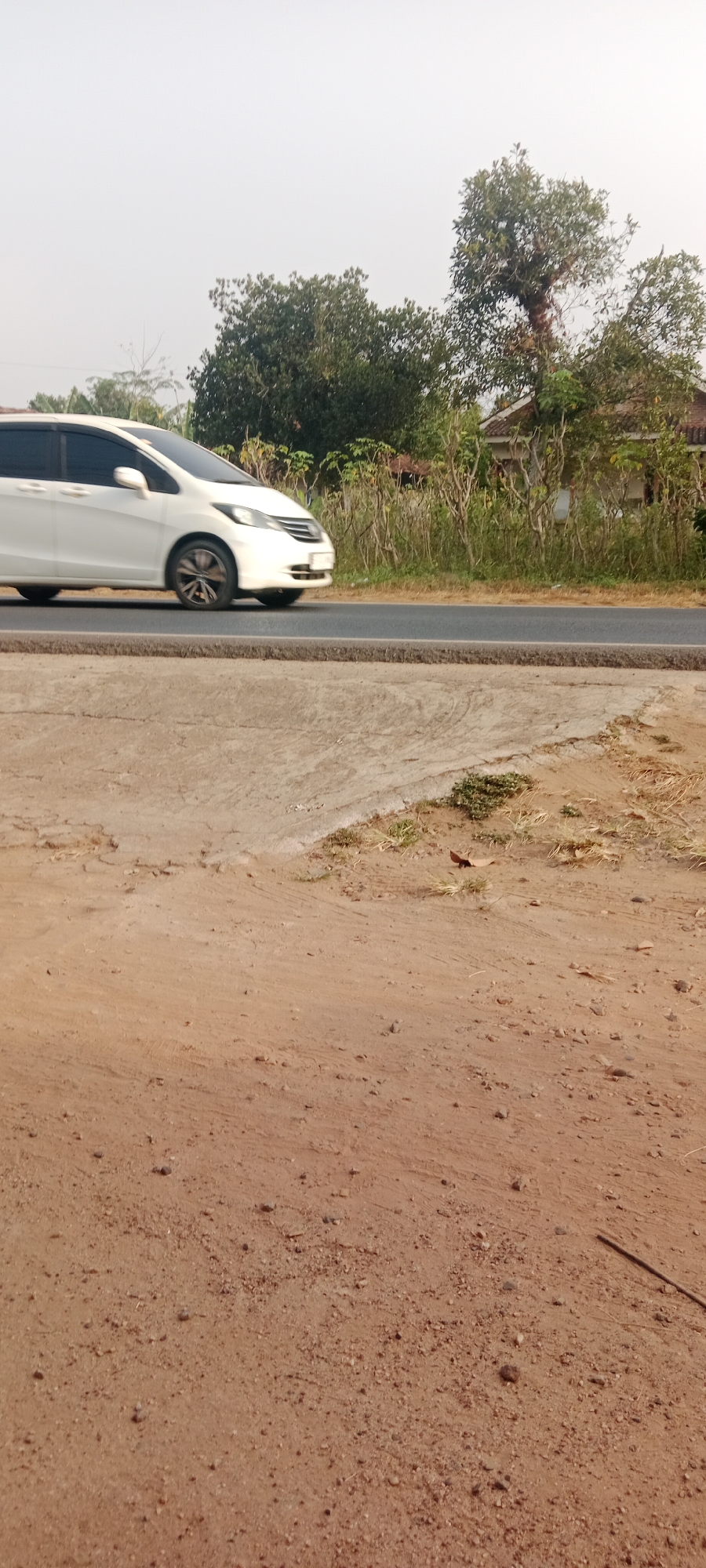 A white car is driving on a road in Gondang Rejo, Indonesia. The car is a minivan, and it is white with tinted windows. It is a sunny day, and the sky is clear. There are trees and bushes on the side of the road. The road is asphalt, and there is a dirt shoulder on the right side of the road. The dirt shoulder is rough and uneven, and there are some small rocks and pebbles on it. There is a concrete curb separating the road from the dirt shoulder. The car is in motion, and its wheels are blurred due to the fast shutter speed. The background is out of focus, but it appears to be a rural area with some houses and vegetation.