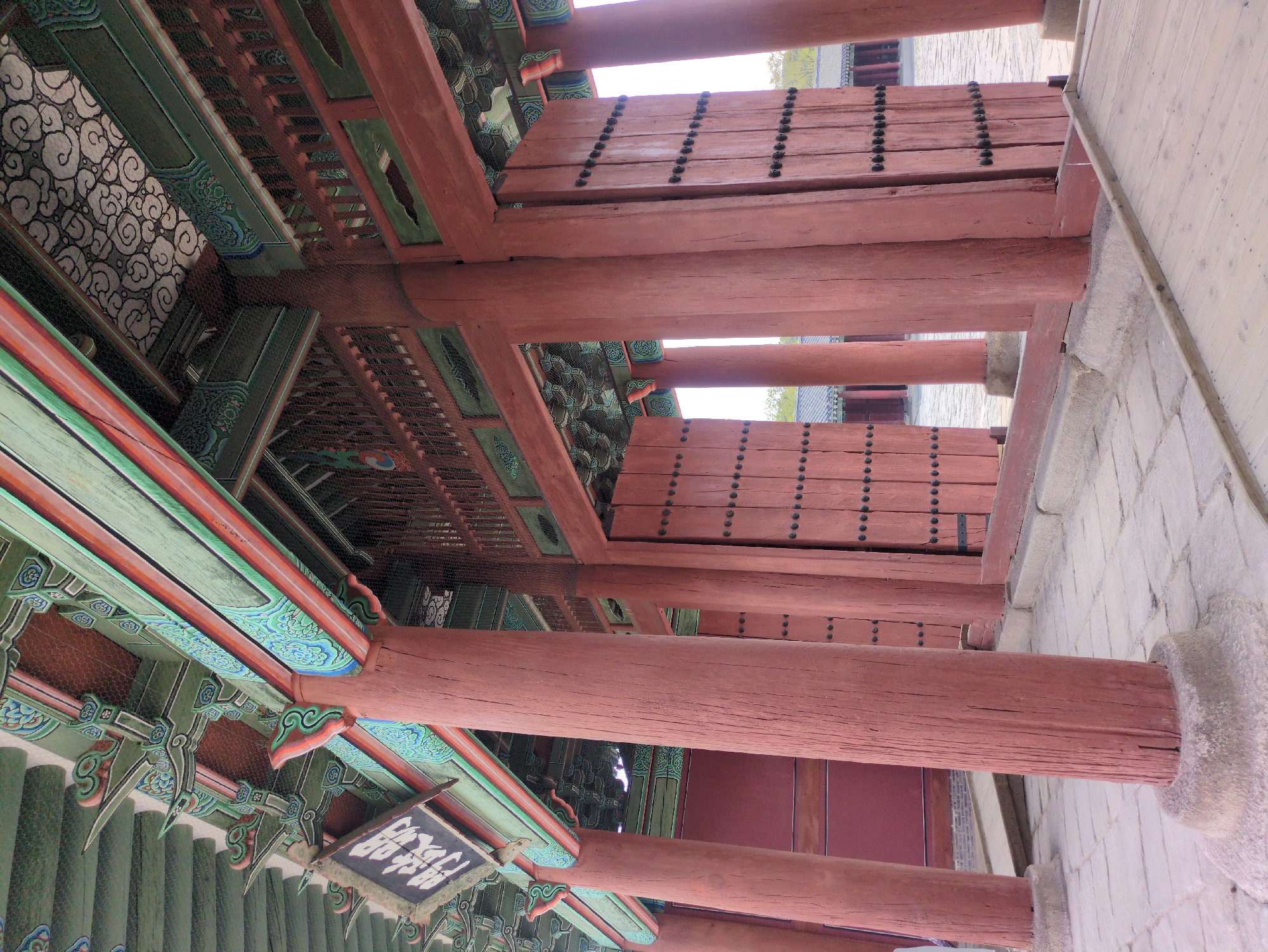 The image depicts the architecture of a traditional Korean building, likely in Seoul, South Korea. Visible are painted wooden columns, beams, and eaves, typical of Korean historical architecture. The coloring is vibrant, featuring green, red, and blue hues with intricate designs. The construction exhibits detailed woodwork and decorative patterns. A sign with Korean script is attached to the structure. The building’s framework, including the roof and support system, is prominent. The orientation of the shot is somewhat tilted, giving a view looking up at the structure from below. A portion of a stone wall is visible on the right side. The environment is outdoors and likely part of a historical site.
