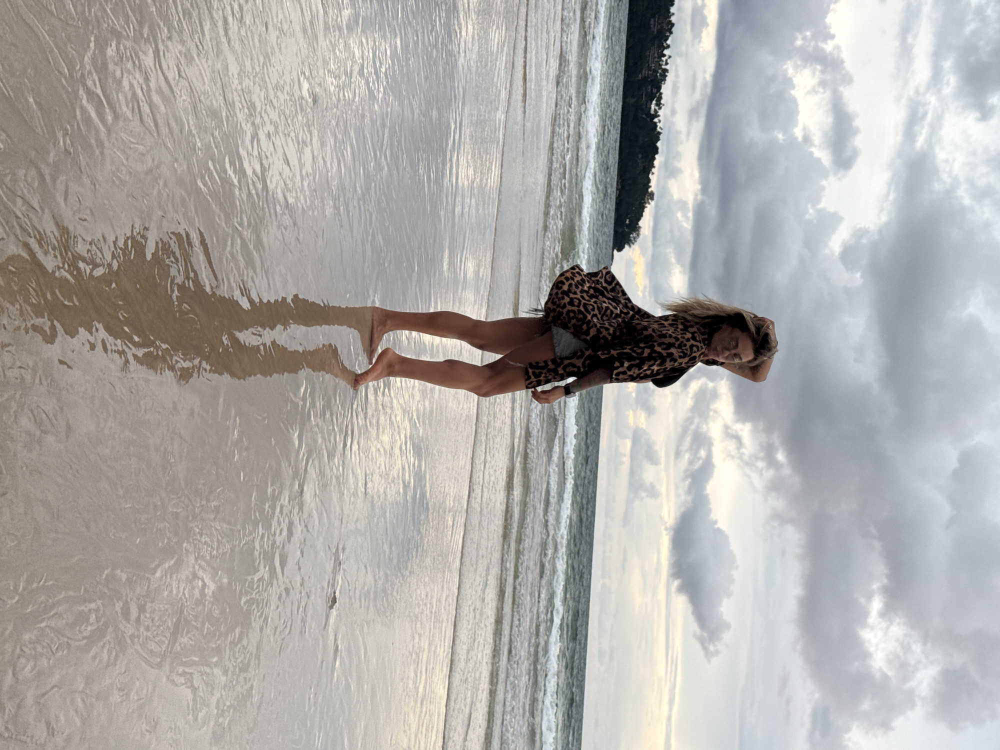 A woman stands on a wet, sandy beach, her reflection shimmering in the shallow water that covers the sand. She is wearing a leopard-print shirt dress over denim shorts. Her hair is blonde and windswept, and she is touching her head with her hand. The waves are gently rolling in on the shore, and the sky is overcast with dramatic, dark clouds. A line of trees can be seen in the distance, suggesting a tropical location. The overall mood is serene and peaceful, with a sense of introspection. The time of day appears to be late afternoon or early evening, given the soft, diffused light and the moody sky. The location is Choeng Thale, Thailand. There is no visible text in the image.