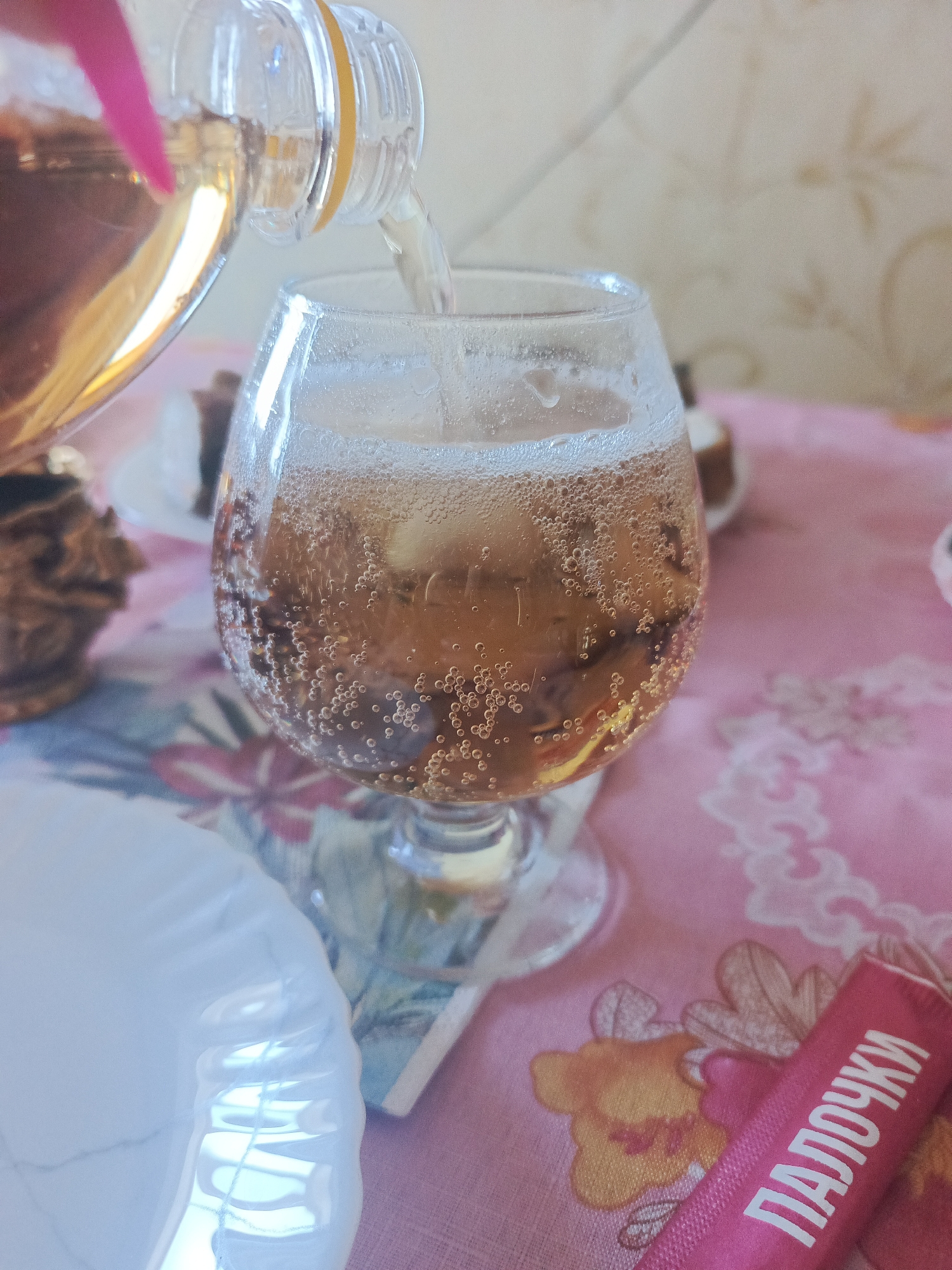 A close-up, eye-level shot shows a bottle of amber liquid being poured into a stemmed glass, creating a fizzy beverage with numerous bubbles. The glass is partially filled, and condensation is visible on its surface. In the background, a patterned tablecloth, likely pink with floral motifs, is visible. To the left of the glass, a decorative, brown object, possibly a small statue or ornament, is partially in view. In the foreground, a white, textured object, possibly a plate or bowl, is present, and a pink wrapper with white text in Cyrillic script, reading "ПАЛОЧКИ", is lying on the tablecloth to the right. The lighting is soft and natural, suggesting an indoor setting. The location is Unknown, Unknown.