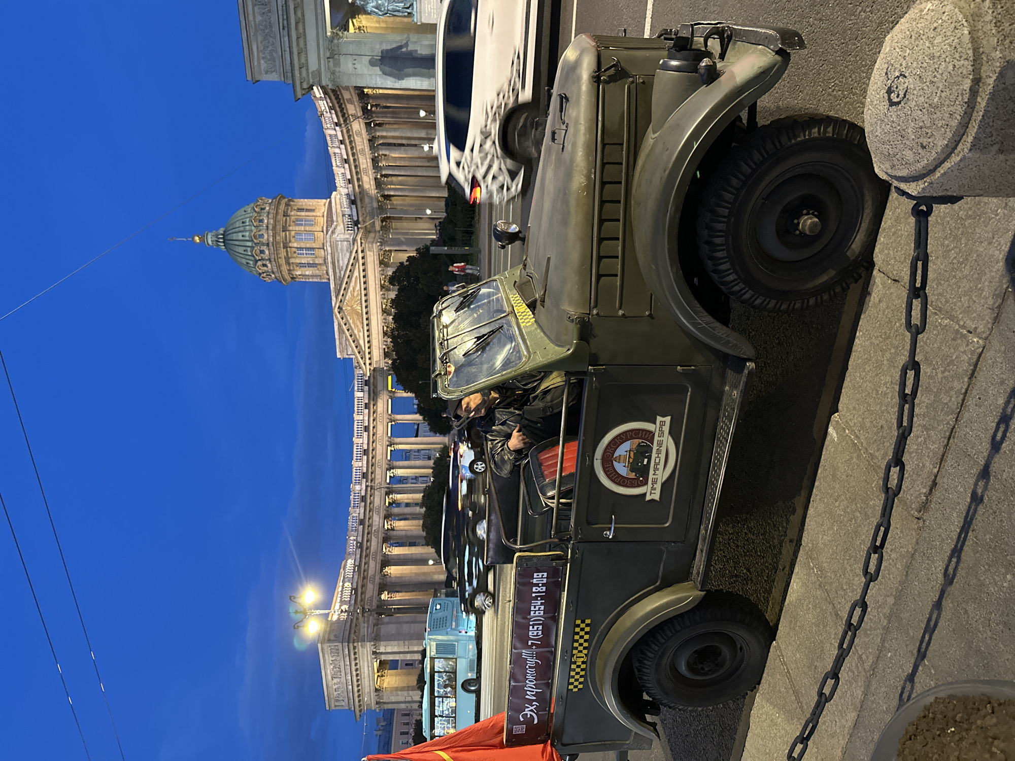 This is a medium shot photograph taken at night in Saint Petersburg, Russia. The main subject is a vintage olive-drab military-style jeep parked on a cobblestone street. A person, wearing a dark jacket, is seated in the driver's seat of the jeep. On the side of the jeep, there are two circular stickers. The top sticker reads "БЗО РНЫЕ ЭКСКУРСИИ TIME MACHINE SPB" and depicts a stylized clock tower and a train. The lower sticker has yellow and black checkered patterns and Russian text that appears to be "Эх, прокачу!!!" followed by a phone number.
In the background, a grand building with a large dome and classical columns is illuminated. This building is characteristic of Saint Petersburg's architecture, likely a historical landmark such as Kazan Cathedral. A light blue bus is visible on the street behind the jeep, and other vehicles are blurred due to motion. The sky is a deep, clear blue, indicating it's twilight or early night. A thick metal chain runs along the sidewalk in the foreground, casting a shadow. The scene suggests a tour or event taking place in a historic urban setting at night.