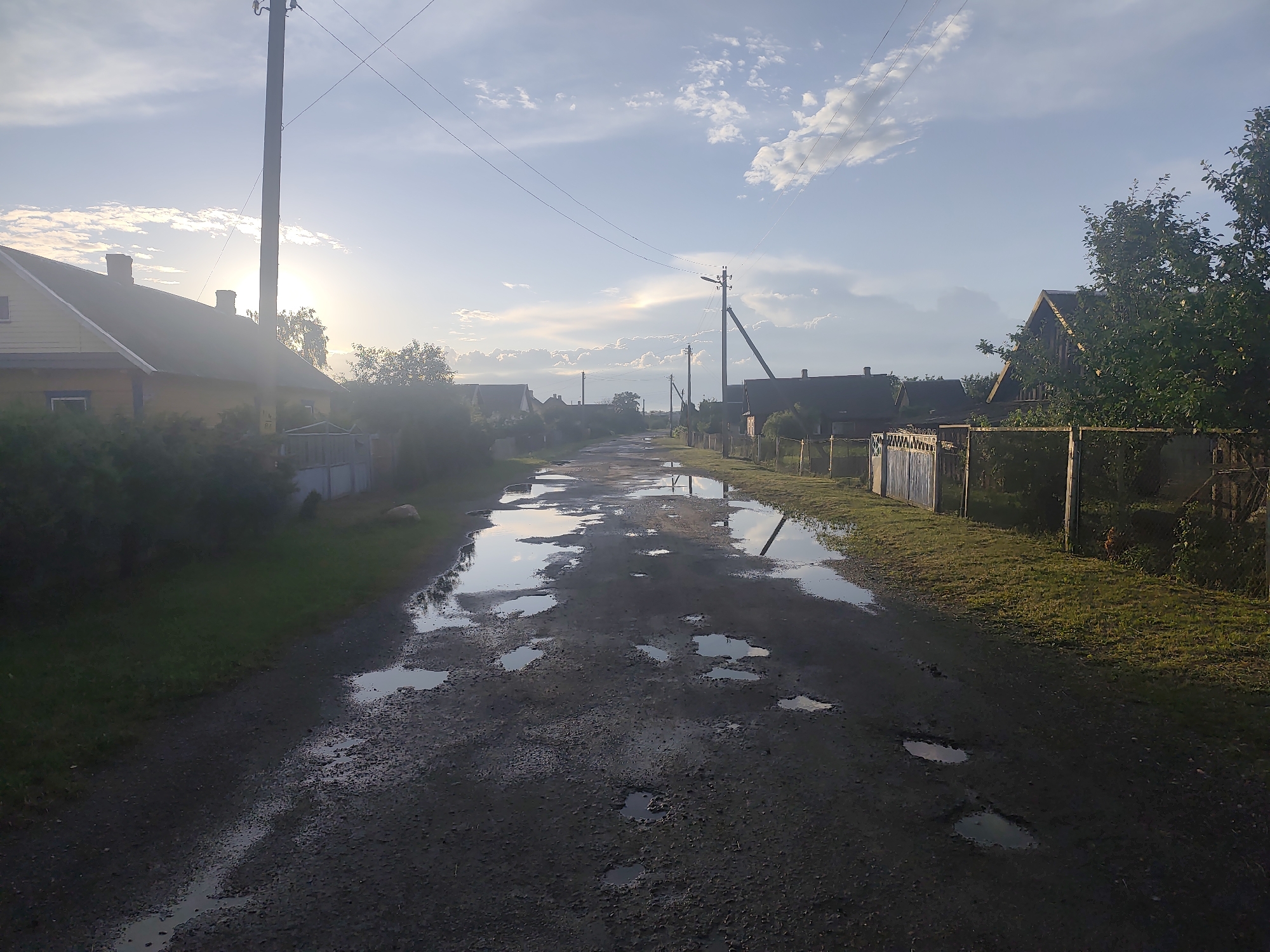 This image captures a rural scene in the Yaglevitski village council, Belarus, featuring a dirt road lined with houses and trees. The road is partially covered in puddles reflecting the cloudy sky, suggesting recent rainfall. Power lines and poles are visible, characteristic of rural infrastructure. The houses appear to be of traditional design, with wooden fences and small yards. The overall impression is of a quiet, serene village setting under a partly sunny sky.