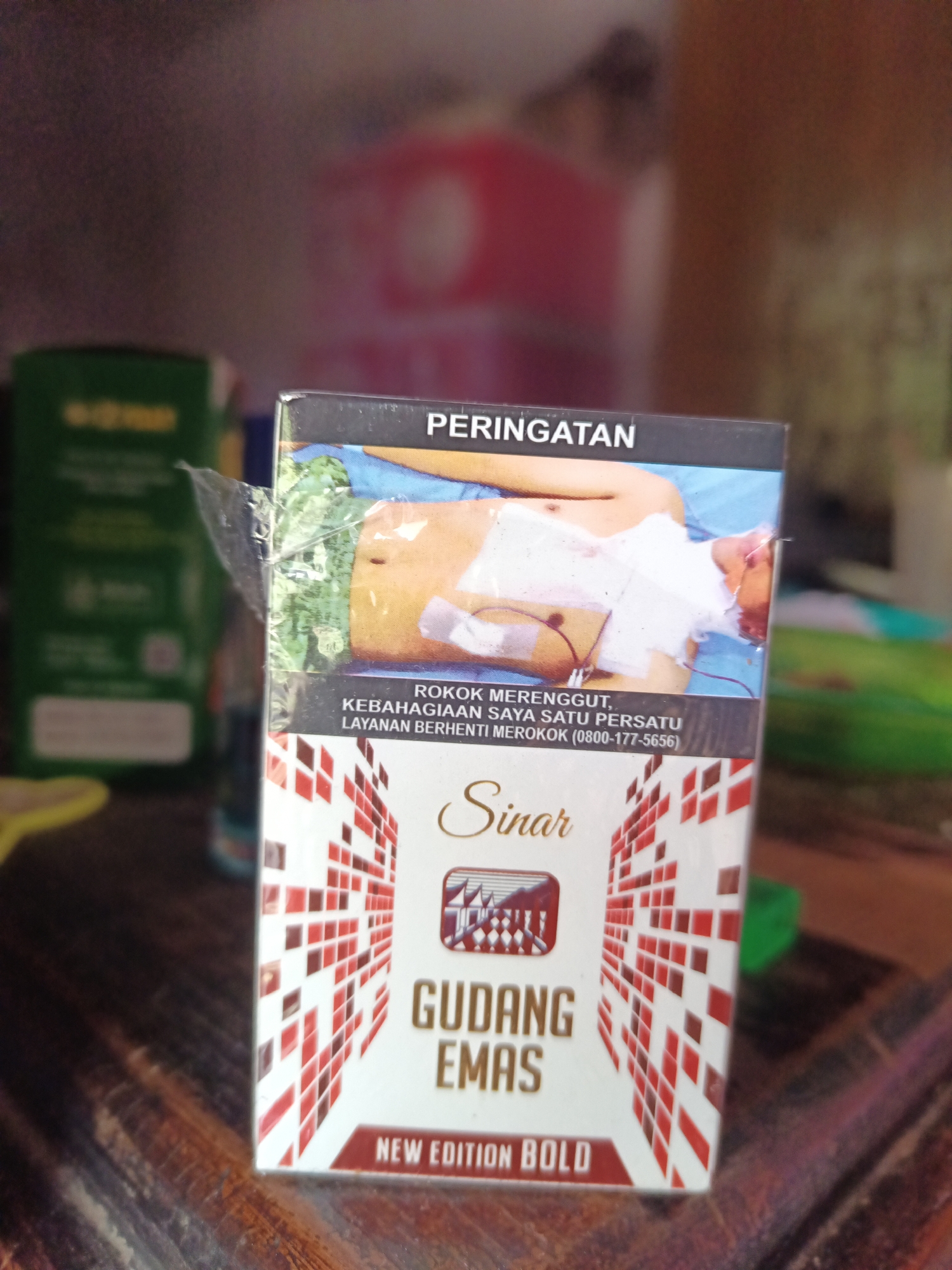 A close-up, eye-level shot shows the front of a cigarette pack. The pack is a light color with red and white geometric patterns on the sides. At the top, there is a black band with white text that reads "PERINGATAN." Below this, a graphic image depicts a person lying down with their chest bandaged. Text below the graphic reads "ROKOK MERENGGUT, KEBAHAGIAAN SAYA SATU PERSATU LAYANAN BERHENTI MEROKOK (0800-177-5656)." In the center of the pack, the brand name "Sinar" is written in script. Below "Sinar" is a crest with a pagoda-like structure. Further down, the words "GUDANG EMAS" are displayed in large, bold letters. At the bottom, in a red rectangle, it says "NEW EDITION BOLD." The background is blurred, but appears to be indoors with hints of other objects and a wooden surface in the foreground. The image was taken in South Jakarta, Indonesia.