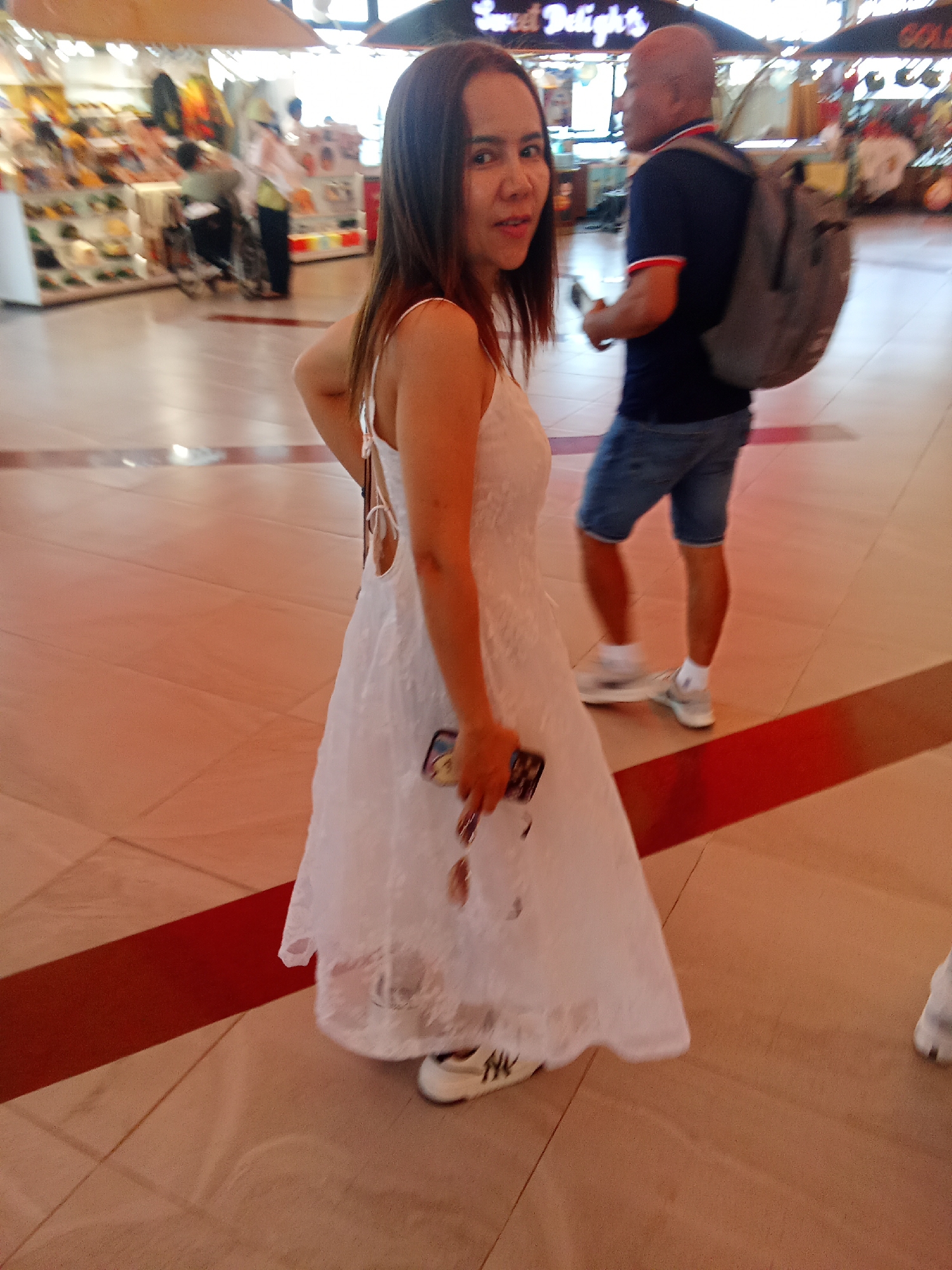 A woman in a white dress is standing in the middle of a brightly lit indoor walkway, possibly a shopping mall, with a blurred background of shops and people. She is looking towards the camera with a slight smile. She is holding a dark object in her right hand, which appears to be a phone. To her right, a man with a shaved head is walking away from the camera, wearing a dark blue t-shirt and denim shorts, carrying a backpack. The floor is tiled with a light brown color, accented by a red stripe running diagonally across the image. The overall lighting suggests a commercial indoor space. This scene is likely located in a shopping center in Đà Nẵng, Vietnam, given the context provided.
