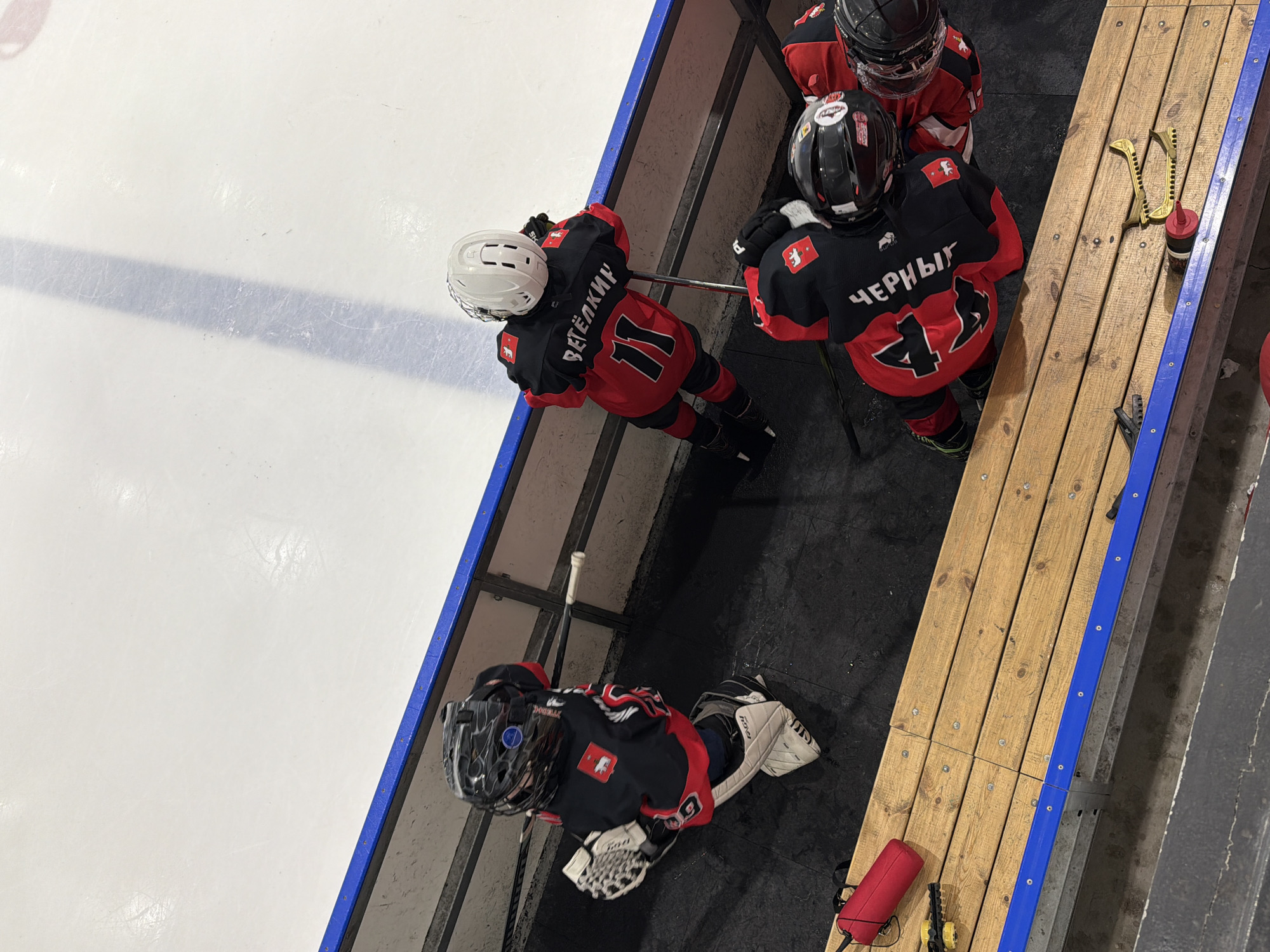 Four young hockey players in red and black jerseys are on the team bench in Votkinsk, Russia. Three are standing, leaning on a metal railing, while one sits on the floor by the bench. The ice rink is visible to the left. There is a wooden bench with equipment, including skate sharpeners and a small red bag.