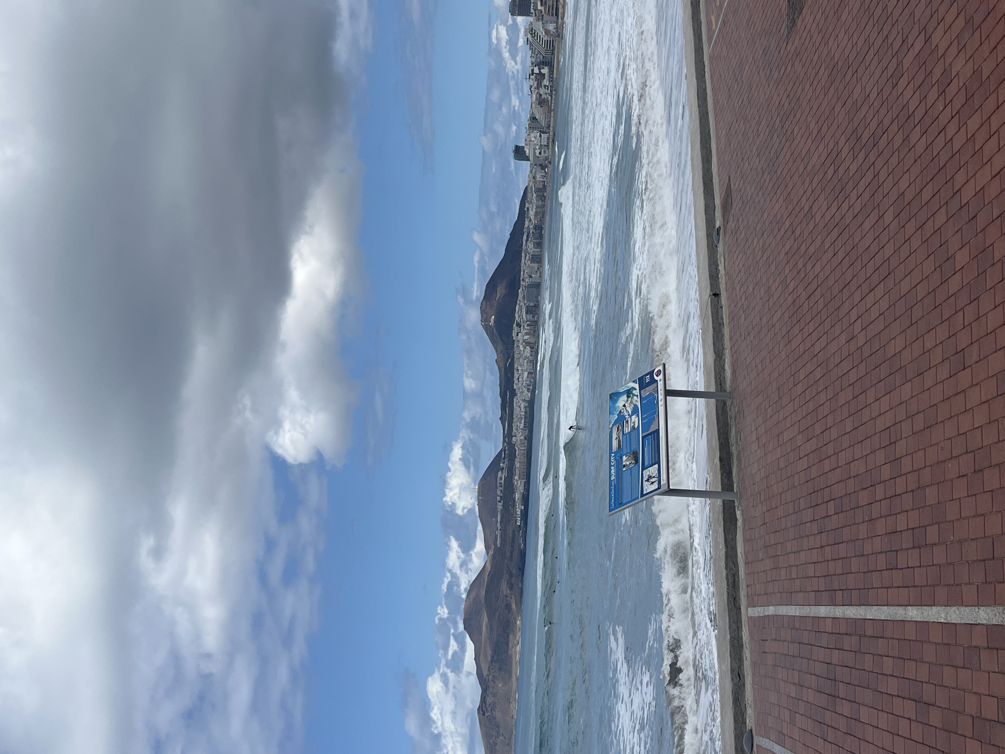 The image captures a coastal scene in Las Palmas De Gran Canaria, Spain, on a bright, partly cloudy day. The perspective is from a promenade looking out over the ocean towards a mountainous coastline and a city.
In the immediate foreground, a wide, red-brown brick-paved promenade runs horizontally, separated from the water by a concrete edge. A blue information sign stands on the promenade, displaying "SURF CITY" prominently in white capital letters. Other smaller text and images are visible on the sign, including what appears to be "La Cicer" and the emergency number "112" in the top right corner.
Beyond the promenade, the ocean is active with breaking white waves. A single surfer is visible on a surfboard in the water, riding a wave, suggesting ongoing surfing activity. In the mid-ground, a long, dark mountainous landmass stretches along the horizon, with a dense urban landscape built into its base and slopes. Numerous buildings, some appearing tall, form the cityscape of Las Palmas.
The sky above is a mix of clear blue patches and scattered, textured white and grey clouds, indicating a partly overcast but bright day. The scene conveys a lively coastal environment, popular for surfing, set against a backdrop of natural beauty and urban development.