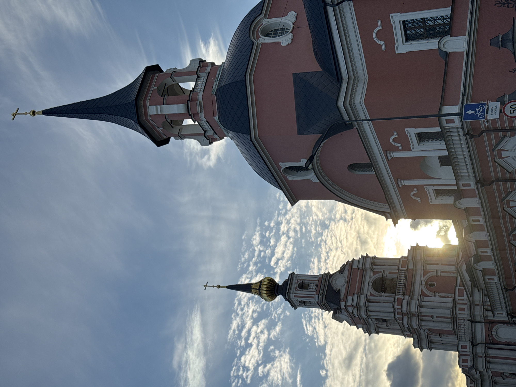 This media file features two ornate church towers against a bright, cloudy sky. The sky is a vibrant blue with streaks of white clouds, suggesting a pleasant day, possibly late afternoon given the warm glow of the sun peeking through. The towers themselves are architecturally detailed, with one being taller and more prominent, topped with a dark, conical spire and a golden cross. The second, shorter tower is closer to the viewer and has a golden, onion-shaped dome also crowned with a cross. Both towers exhibit intricate stonework, with white decorative elements contrasting against a salmon-pink facade.
Visible architectural details include arched windows, pilasters, and ornate cornices. The larger tower shows a section of the main church building, which is also pink with dark blue tiled roofing. A street sign with a bicycle symbol and Cyrillic text is visible on a pole attached to the building. The text on the sign reads "отмена - / «исключ»". A circular sign with the number "50" is also visible. The perspective is upward-looking, emphasizing the grandeur of the religious structures. The scene evokes a sense of historical significance and architectural beauty, set against a dynamic sky. The sunlight casts a warm, slightly hazy light, highlighting the textures and forms of the buildings.